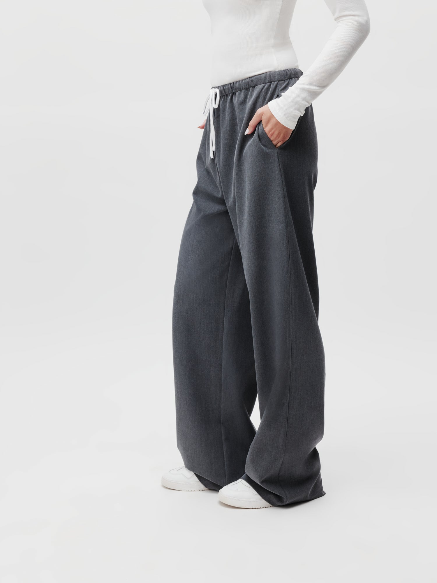 Eine Person steht selbstbewusst da und präsentiert ihr Outfit aus Kim Pants von LeGer by Lena Gercke in Größe 38 und einem langärmeligen weißen Hemd. Die weiten grauen Hosen haben einen Kordelzug in der Taille und vervollständigen den Look mit weißen Sneakers auf einem schlichten weißen Hintergrund.