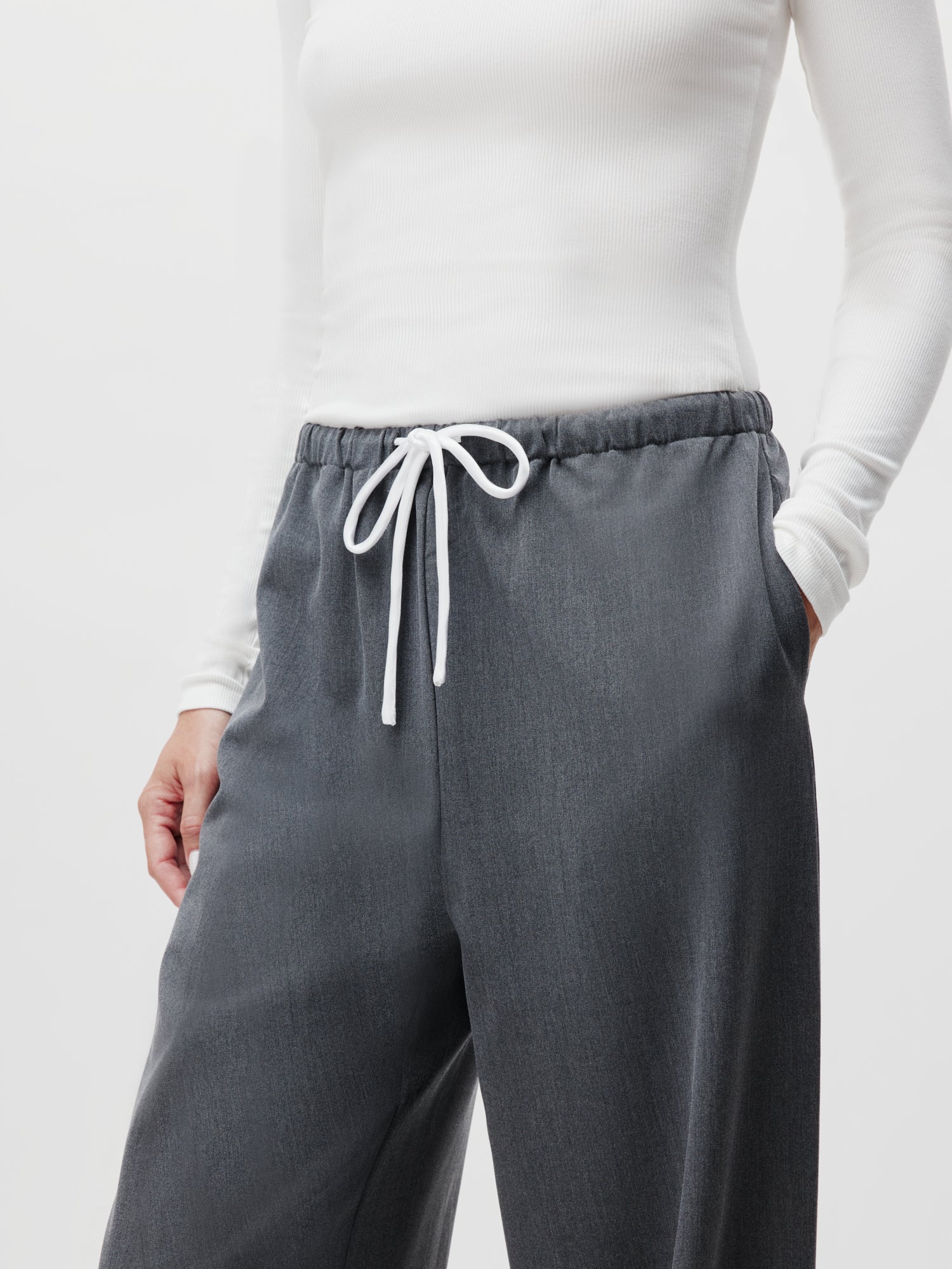 Ein Model trägt ein langärmliges weißes Top und Kim Pants von LeGer by Lena Gercke, Größe 38, und hat eine Hand in der Tasche. Der Fokus liegt auf dem Outfit, das die Textur und Passform der Kleidung vor einem schlichten Hintergrund hervorhebt.