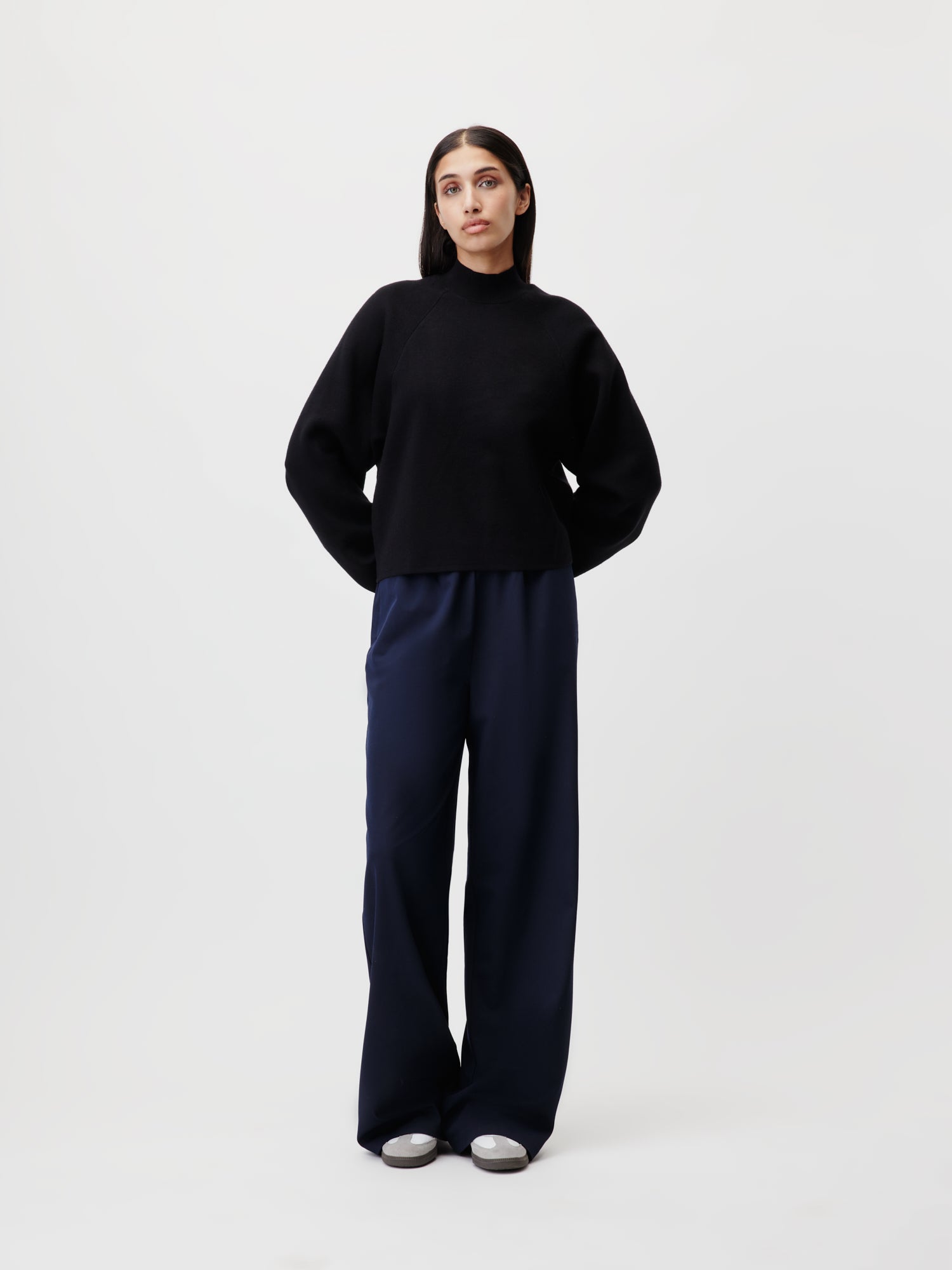 Eine Person mit langen dunklen Haaren steht in einer minimalistischen Umgebung und präsentiert das Modell Größe 36. Sie trägt einen schwarzen Pullover, weit geschnittene marineblaue Kim Pants von LeGer by Lena Gercke von Länge: Lang und weiße Schuhe. Der schlichte weiße Hintergrund betont ihre entspannte Pose und hebt die Produktinformationen mühelos hervor.