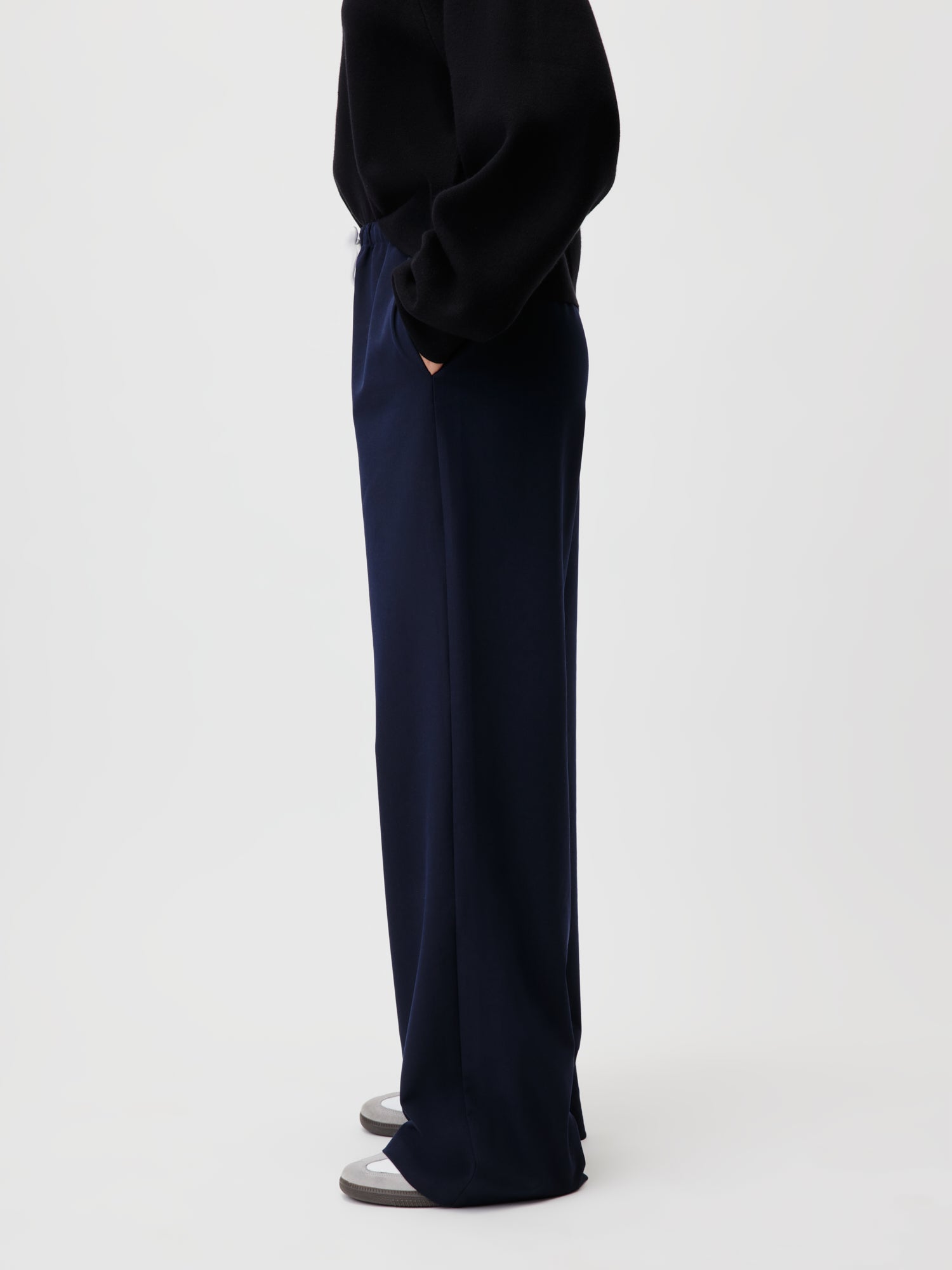 Vor einem schlichten hellen Hintergrund präsentiert eine Person in einem schwarzen Oberteil und der Kim Pants in Blau von LeGer by Lena Gercke in Modelgröße 36 ihren Stil. Sie ist zur Seite gewandt und hat die Hände in den Taschen, so dass die Unterseite ihrer grauen Schuhe zu sehen ist. Die Hose ist im Lang/Maxi-Stil geschnitten.