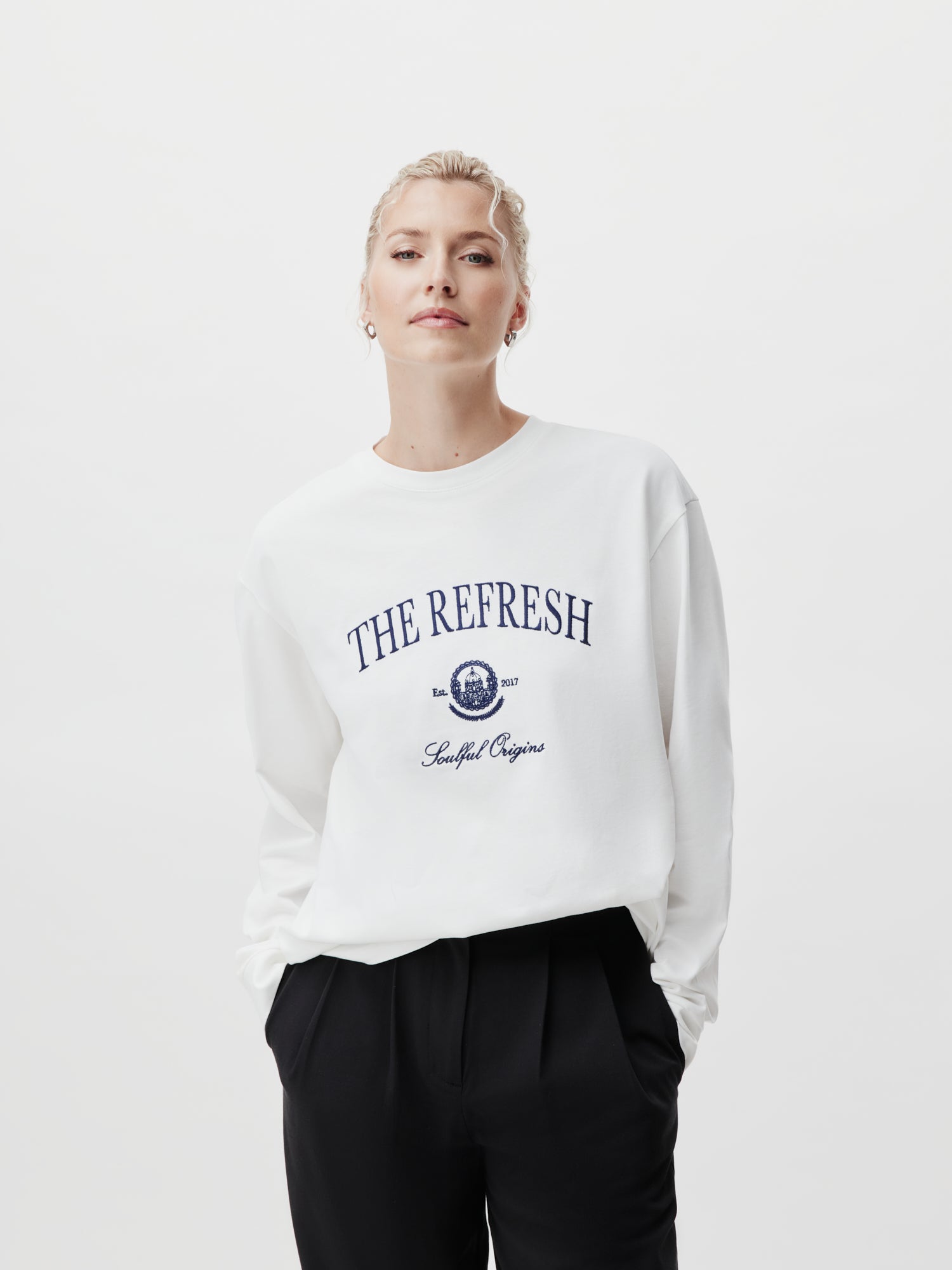 Eine Person mit blondem Haar trägt ein weißes Valeska-Langarmshirt von LeGer by Lena Gercke, auf dem in marineblauem Text „The Refresh“, „Est. 2021“ und „Seoullif Origin“ stehen. Ihre Hände liegen in den Taschen einer schwarzen Hose in normaler Länge vor einem weißen Hintergrund.