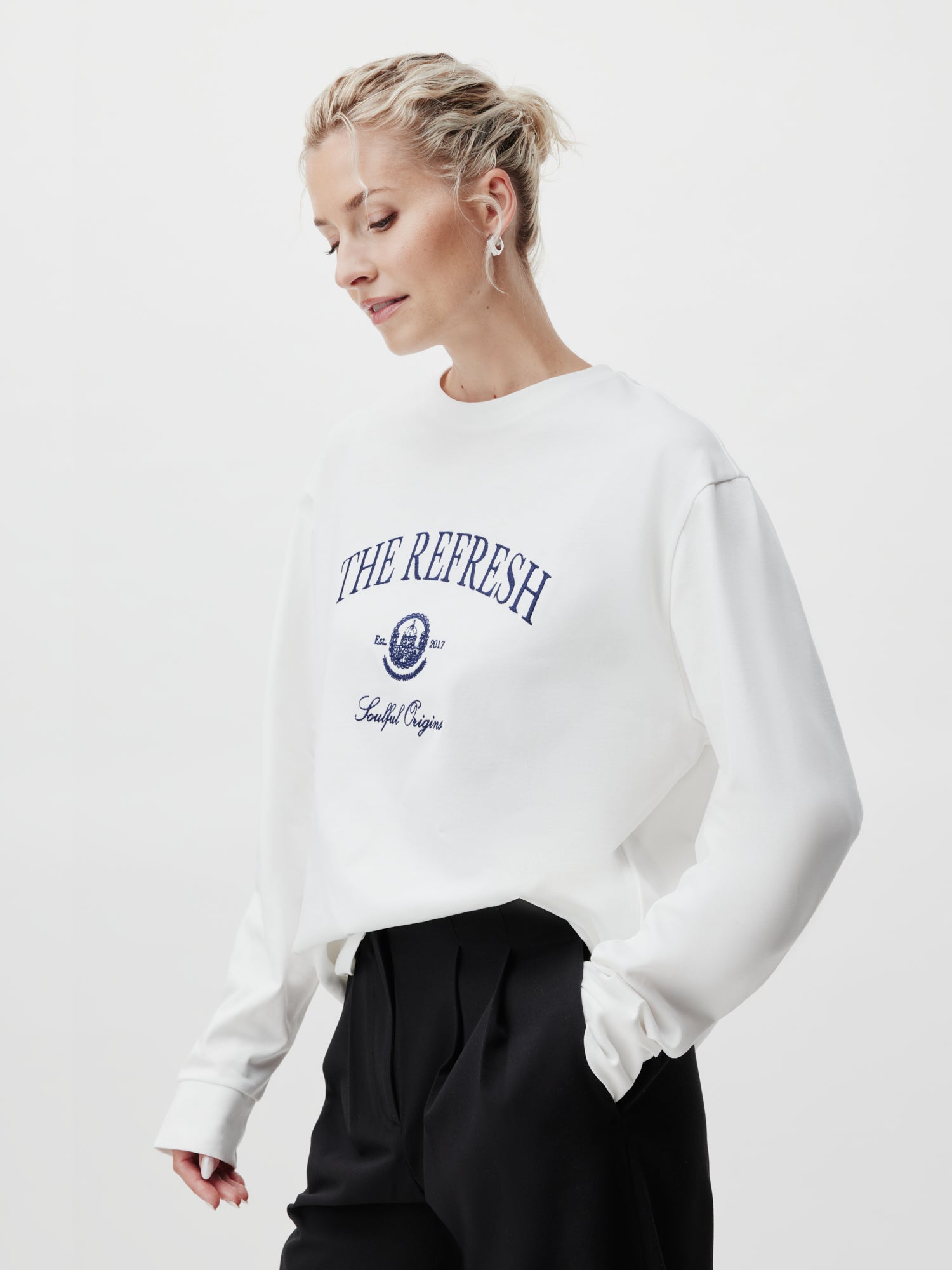 Eine Person mit kurzen blonden Haaren steht vor einem weißen Hintergrund und trägt das Valeska Longsleeve von LeGer by Lena Gercke. Das weiße Sweatshirt hat einen blauen Text mit der Aufschrift „THE REFRESH“ und ist normal lang mit langen Ärmeln. Sie kombinieren es mit einer schwarzen Hose und Ohrringen als Accessoires und behalten so einen heiteren Ausdruck.