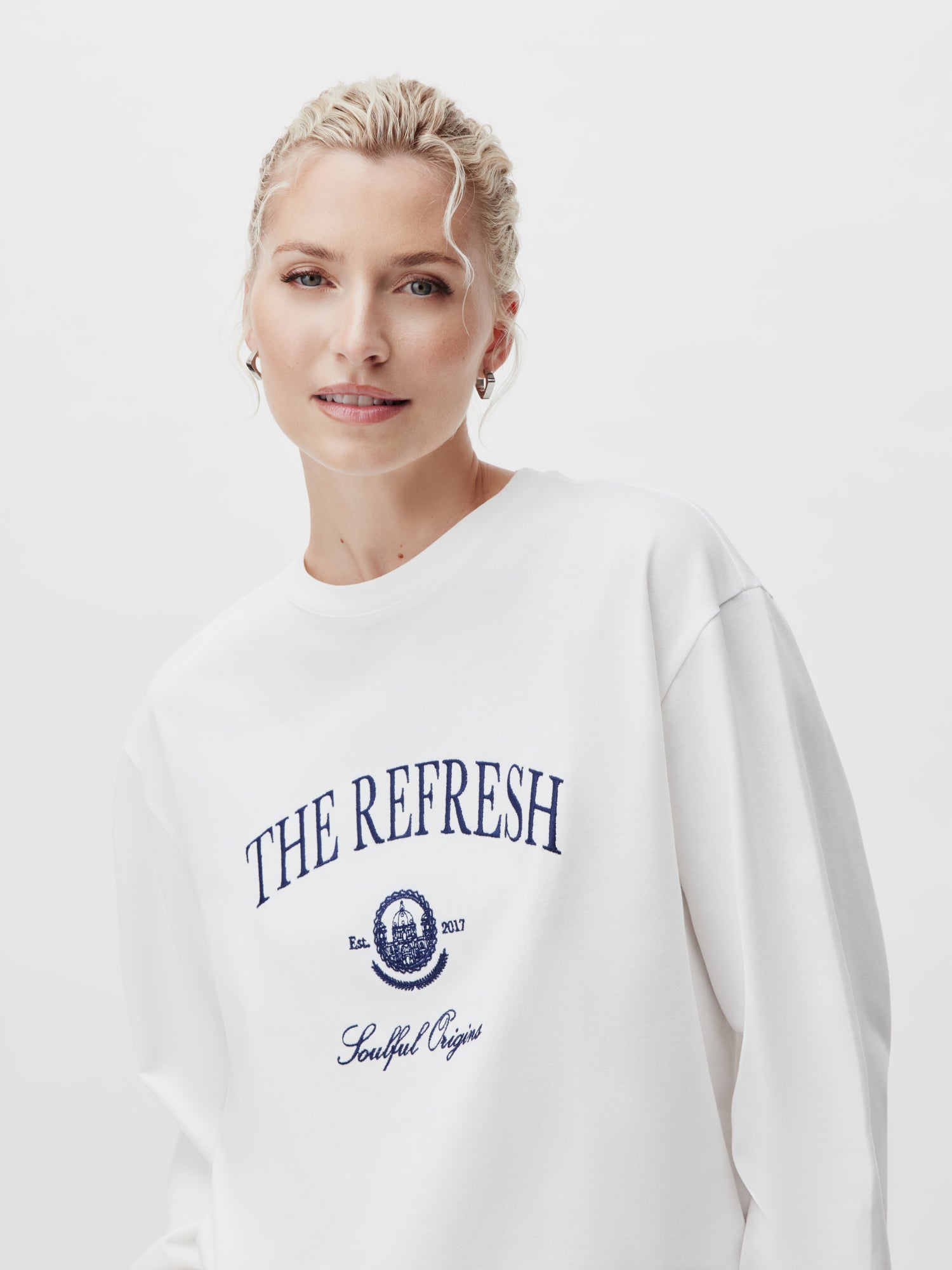 Eine Person mit hellblondem Haar trägt ein weißes Valeska Longsleeve von LeGer by Lena Gercke und trägt in großen marineblauen Buchstaben die Aufschrift „THE REFRESH“. Das Design enthält subtile Details wie „Est. 2017“ und „Soulful Origins“, die die gefühlvollen Ursprünge des Produkts vor einem schlichten weißen Hintergrund hervorheben.