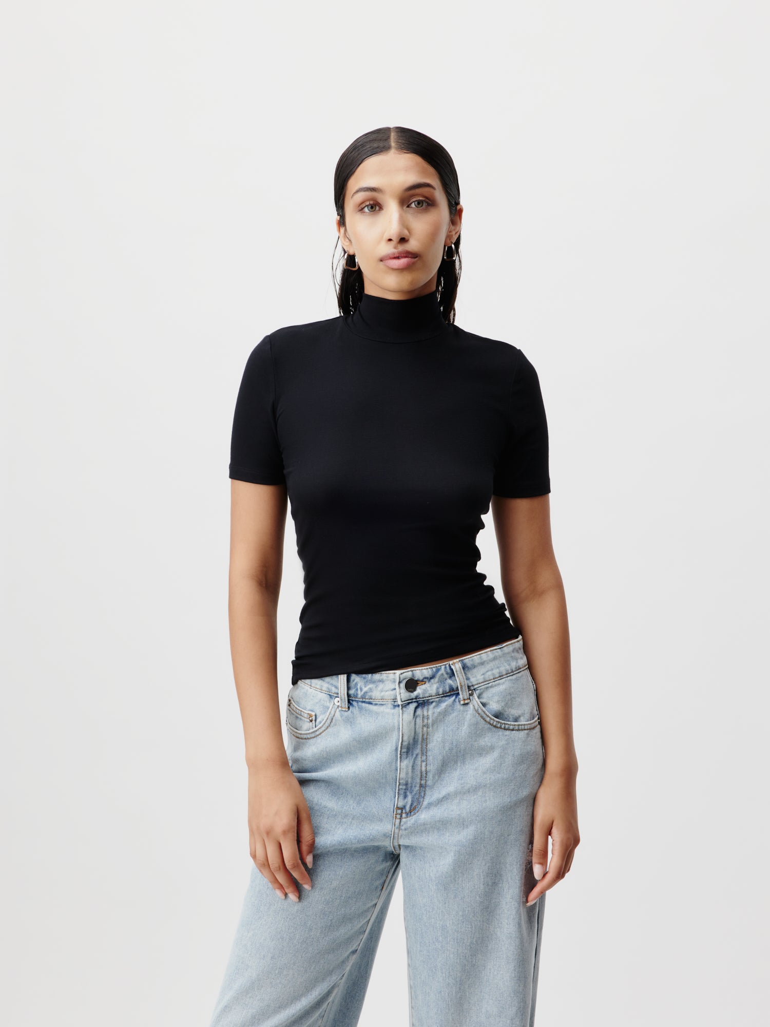 Eine Person mit langen dunklen Haaren steht vor einem schlichten Hintergrund und trägt das Larina T-Shirt von LeGer by Lena Gercke in Schwarz und hellblaue Jeans. Das Outfit schmeichelt der Figur und betont ihre Silhouette. Die Pose ist entspannt, ein Arm ist leicht angewinkelt, was den Viertelarm-Stil des Rollkragenpullovers hervorhebt.