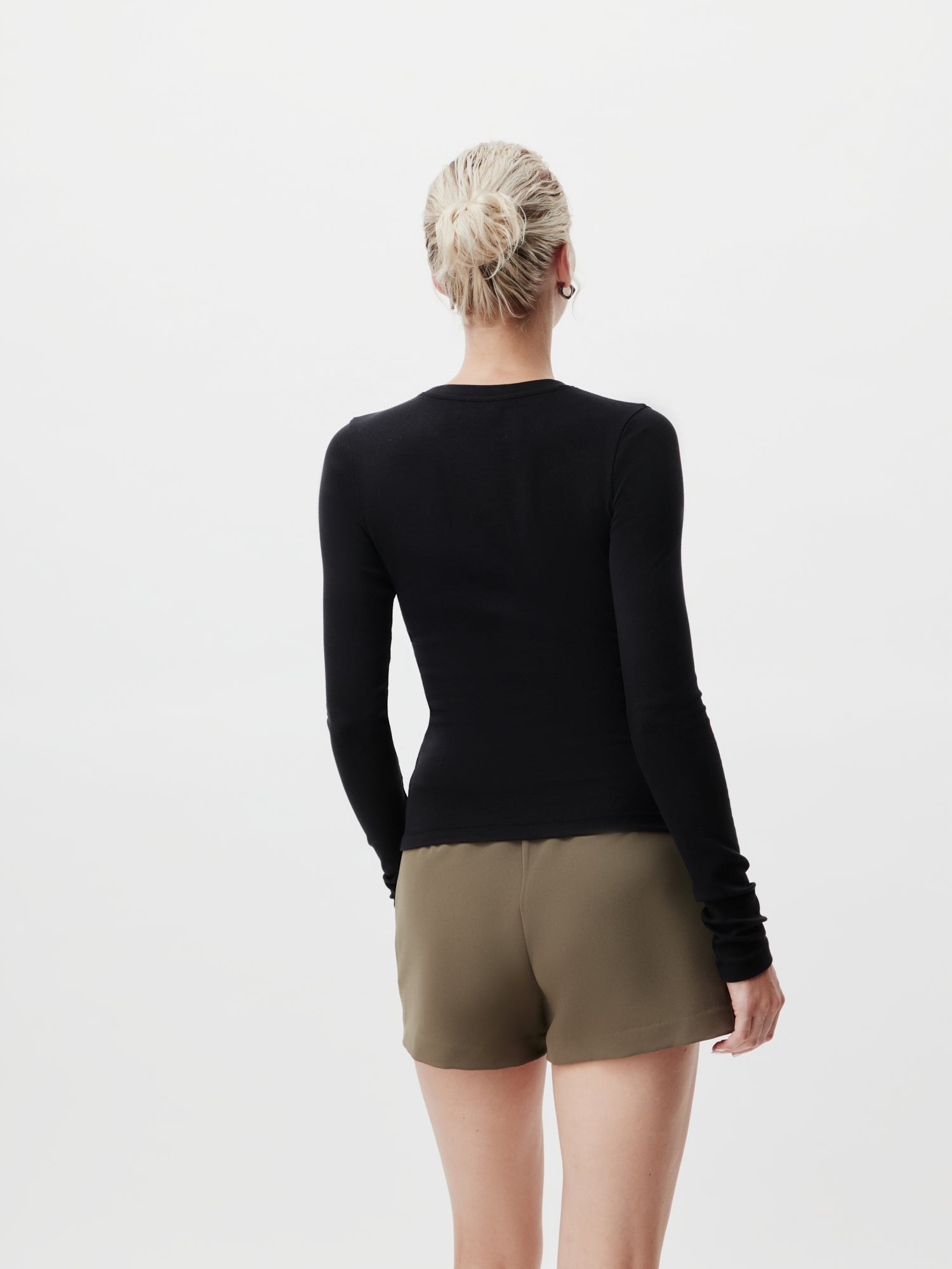 Eine Person mit blonden Haaren im Dutt blickt abgewandt und trägt das Leah Longsleeve von LeGer by Lena Gercke in schwarzen und olivgrünen Shorts. Der Hintergrund ist schlicht weiß.