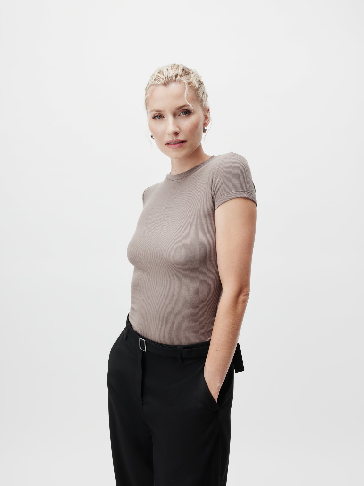 Eine Person mit zurückgebundenem hellem Haar trägt ein schmal geschnittenes, graues Cindy-T-Shirt von LeGer by Lena Gercke mit Viertelarm-Ärmeln. Sie kombiniert es mit schwarzen Hosen normaler Länge und steht vor einem schlichten weißen Hintergrund, die Hände in den Taschen, und blickt in die Kamera.