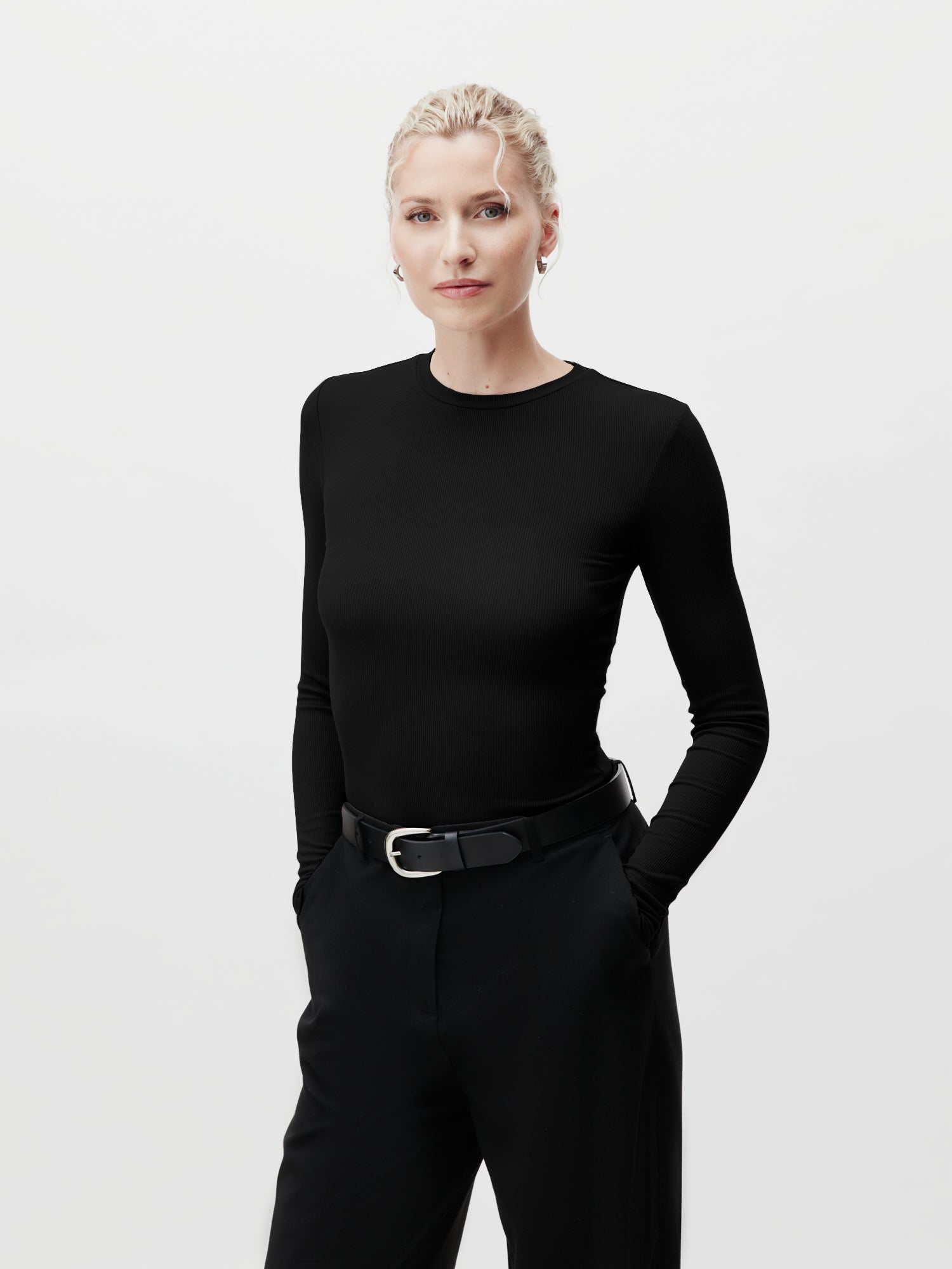 Vor einem schlichten Hintergrund steht eine Person mit hellem Haar, sie trägt ein schwarzes Cinzia Longsleeve von LeGer by Lena Gercke und eine schwarze Hose normaler Länge mit Gürtel. Ihre Hände stecken in den Taschen und zeigen eine Passform Schmal.