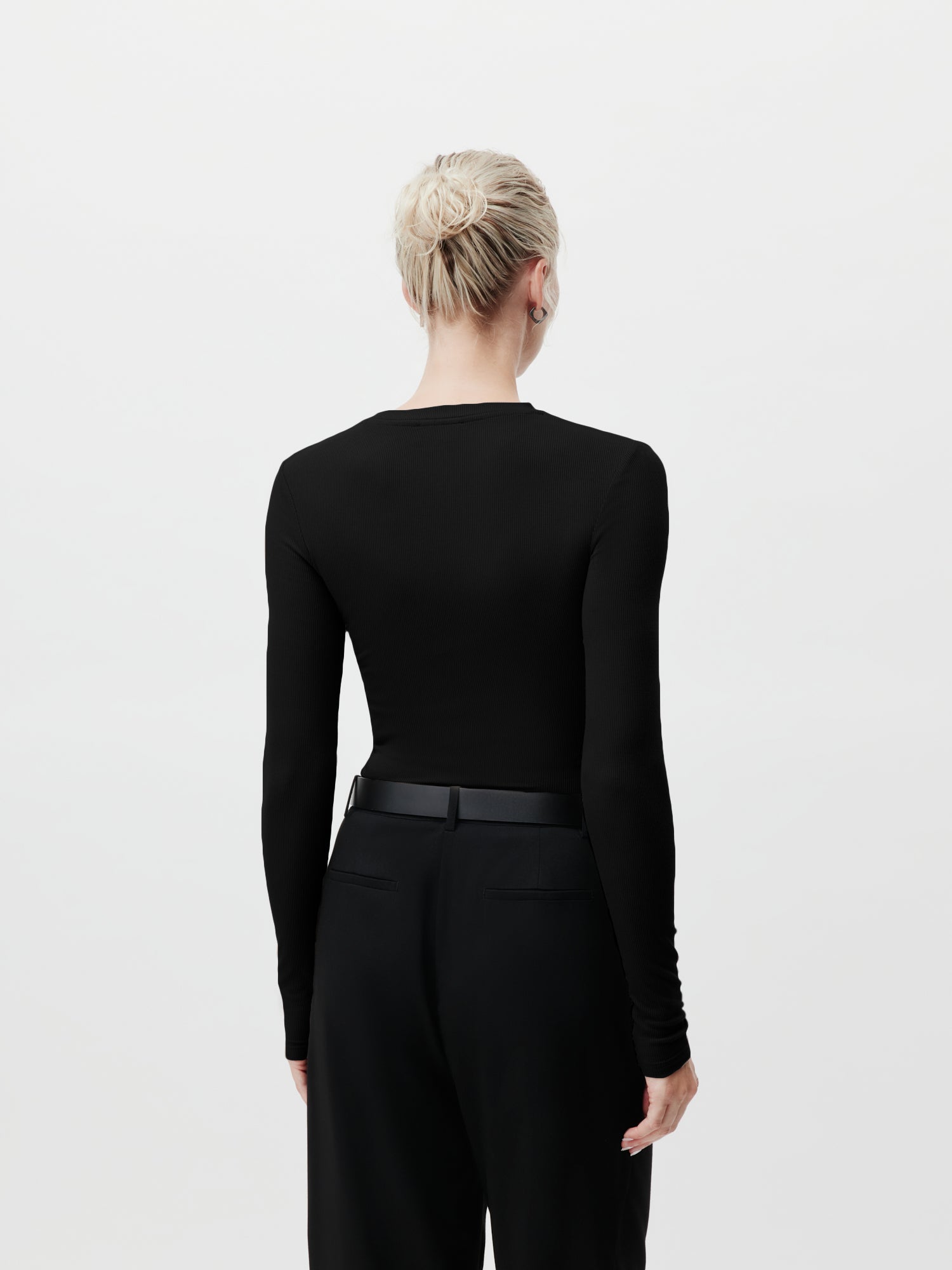 Zu sehen ist eine Person mit blonden Haaren im Dutt von hinten. Sie trägt das Cinzia Longsleeve von LeGer by Lena Gercke, welches durch seine schmale Passform hervorsticht, sowie eine hoch taillierte schwarze Hose. Der Hintergrund ist schlicht weiß.