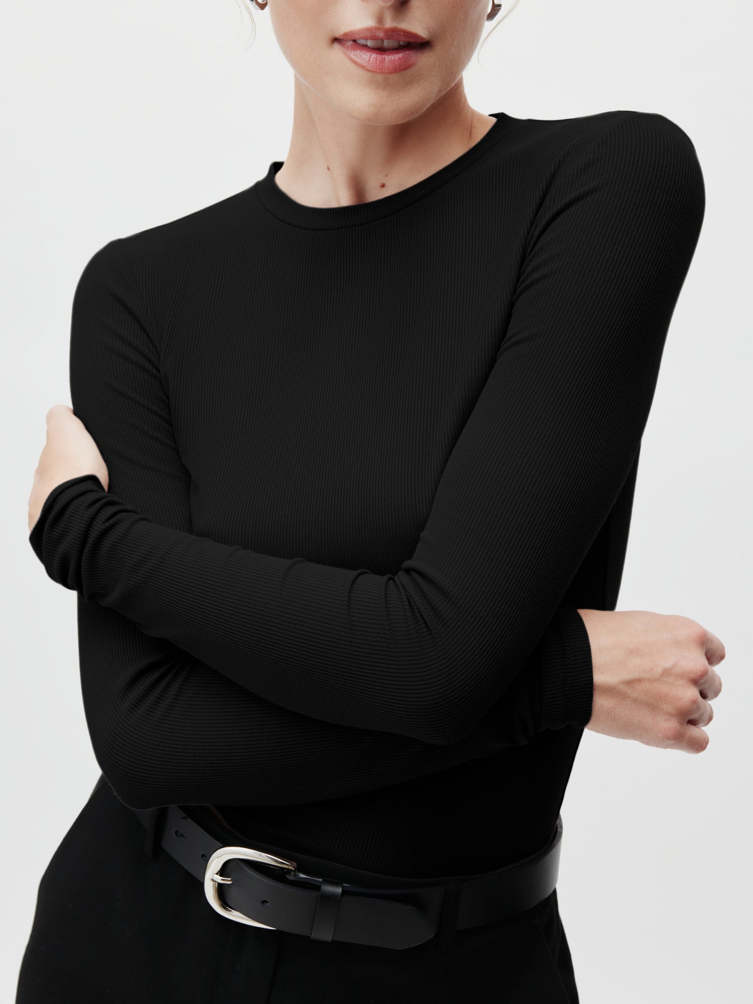 Eine Person trägt das Cinzia Longsleeve von LeGer by Lena Gercke, das sich durch seine schmale Passform und die langen Ärmel auszeichnet. Sie hat die Arme mit neutralem Gesichtsausdruck verschränkt und trägt dazu eine schwarze Hose mit Gürtel in Standardlänge. Der Hintergrund ist schlicht und hell.