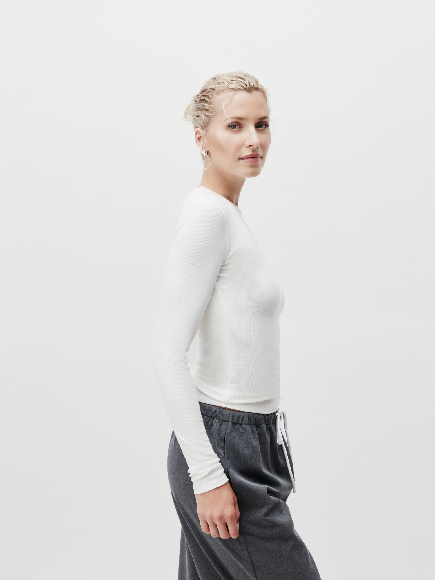 Vor einem schlichten Hintergrund steht eine Person mit kurzen hellen Haaren, trägt ein Cinzia Longsleeve Top von LeGer by Lena Gercke in Weiß und eine schmale graue Hose in Standardlänge. Sie blickt mit neutralem Gesichtsausdruck zur Seite.