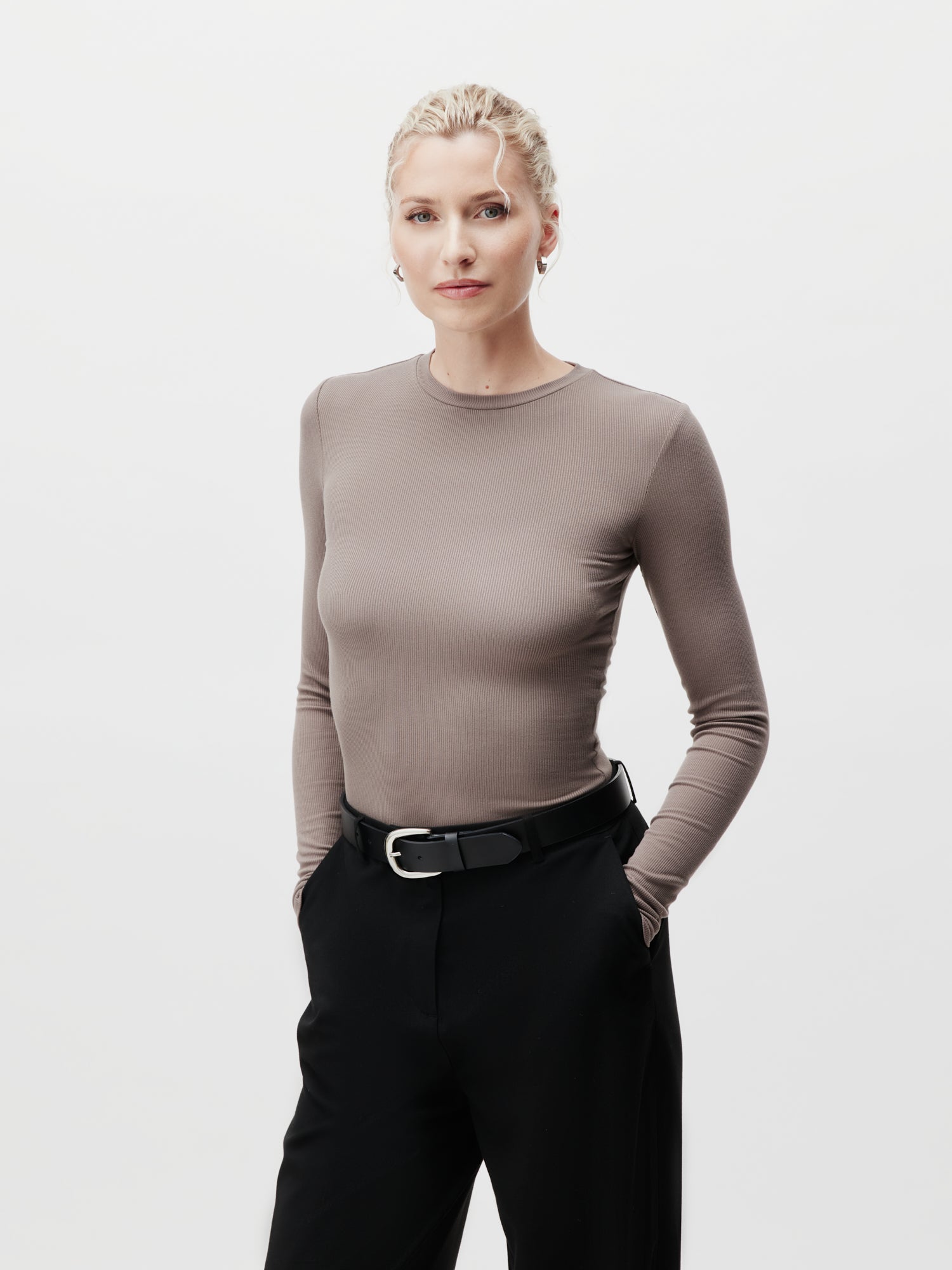 Eine Frau mit kurzen blonden Haaren, gekleidet in ein taupefarbenes Cinzia Longsleeve von LeGer by Lena Gercke im Slim Fit und eine schwarze Hose in Größe 36, steht selbstbewusst mit den Händen in den Taschen vor einem schlichten weißen Hintergrund.