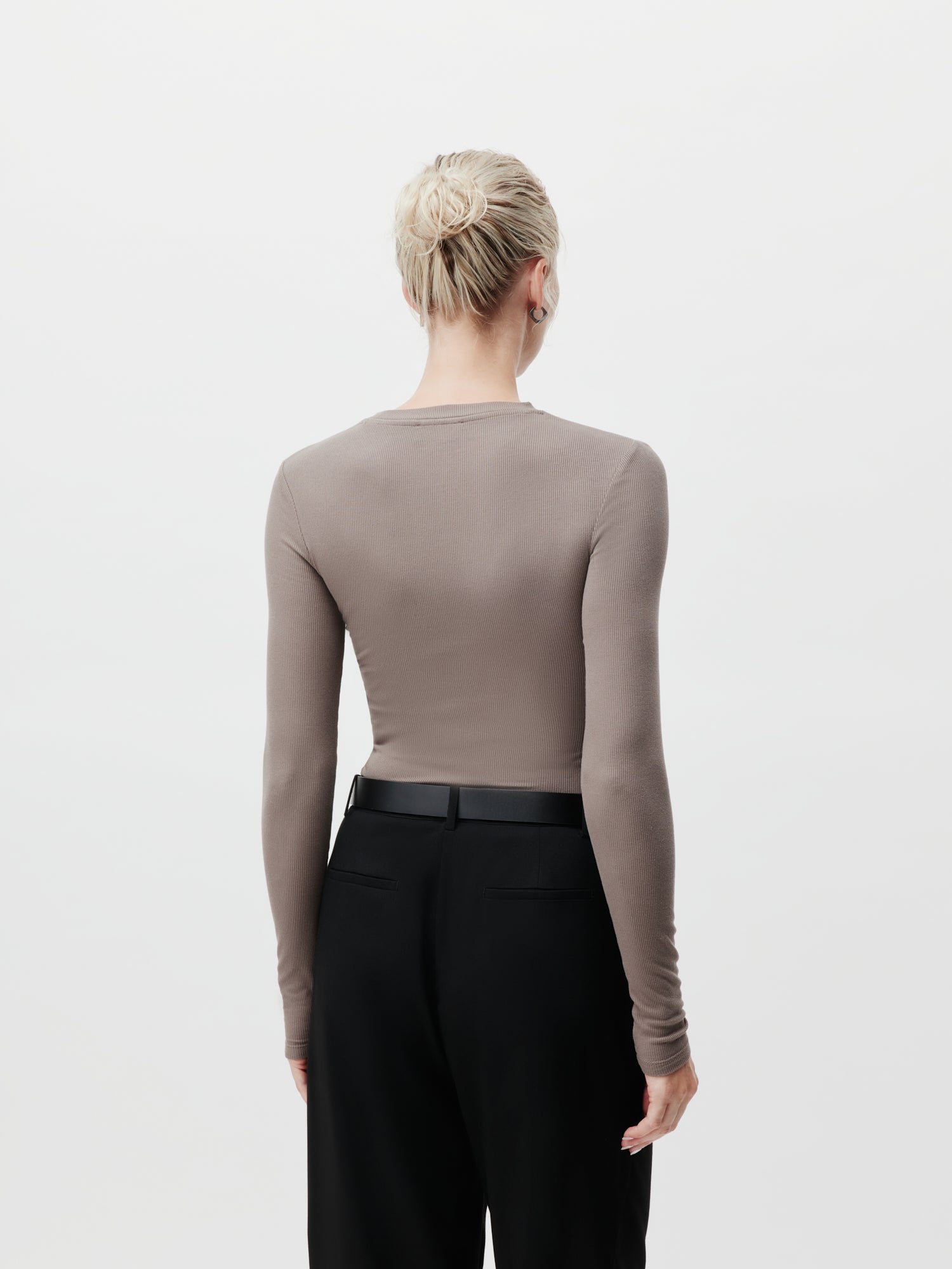 Von hinten ist eine Person mit blonden Haaren zu sehen, die zu einem Dutt gebunden ist. Sie trägt ein LeGer by Lena Gercke Cinzia Longsleeve in Taupe und eine hoch taillierte schwarze Hose in Größe 36. Der Hintergrund ist schlicht weiß.