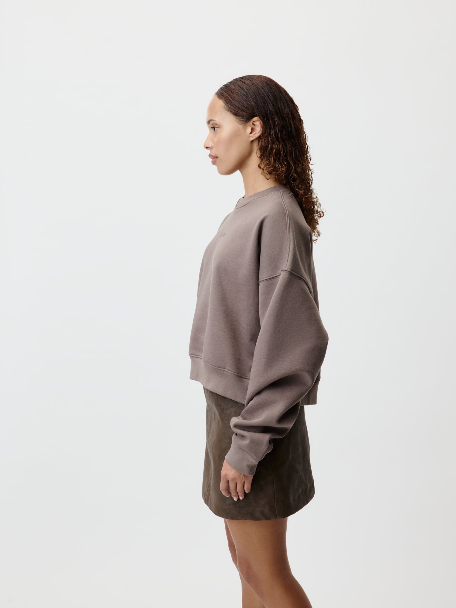 Die Frau ist im Profil zu sehen, bekleidet mit einem taupefarbenen Antonietta-Pullover von LeGer by Lena Gercke mit bequemer Passform und langen Ärmeln, kombiniert mit einem olivfarbenen Rock, und steht vor einem schlichten weißen Hintergrund.