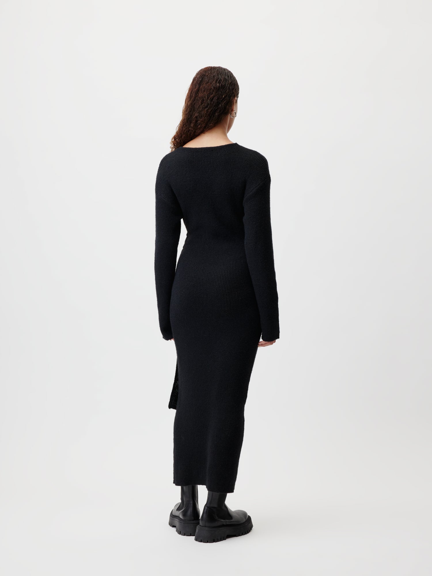 Eine Rückansicht einer Person, die das Bryna-Kleid von LeGer by Lena Gercke trägt. Das elegante, langärmlige schwarze Design mit Seitenschlitz ist besonders schön anzusehen. Das Outfit wird mit schwarzen Stiefeln kombiniert und die Person zeigt ihr langes lockiges Haar, während sie vor einem schlichten, hellen Hintergrund steht.