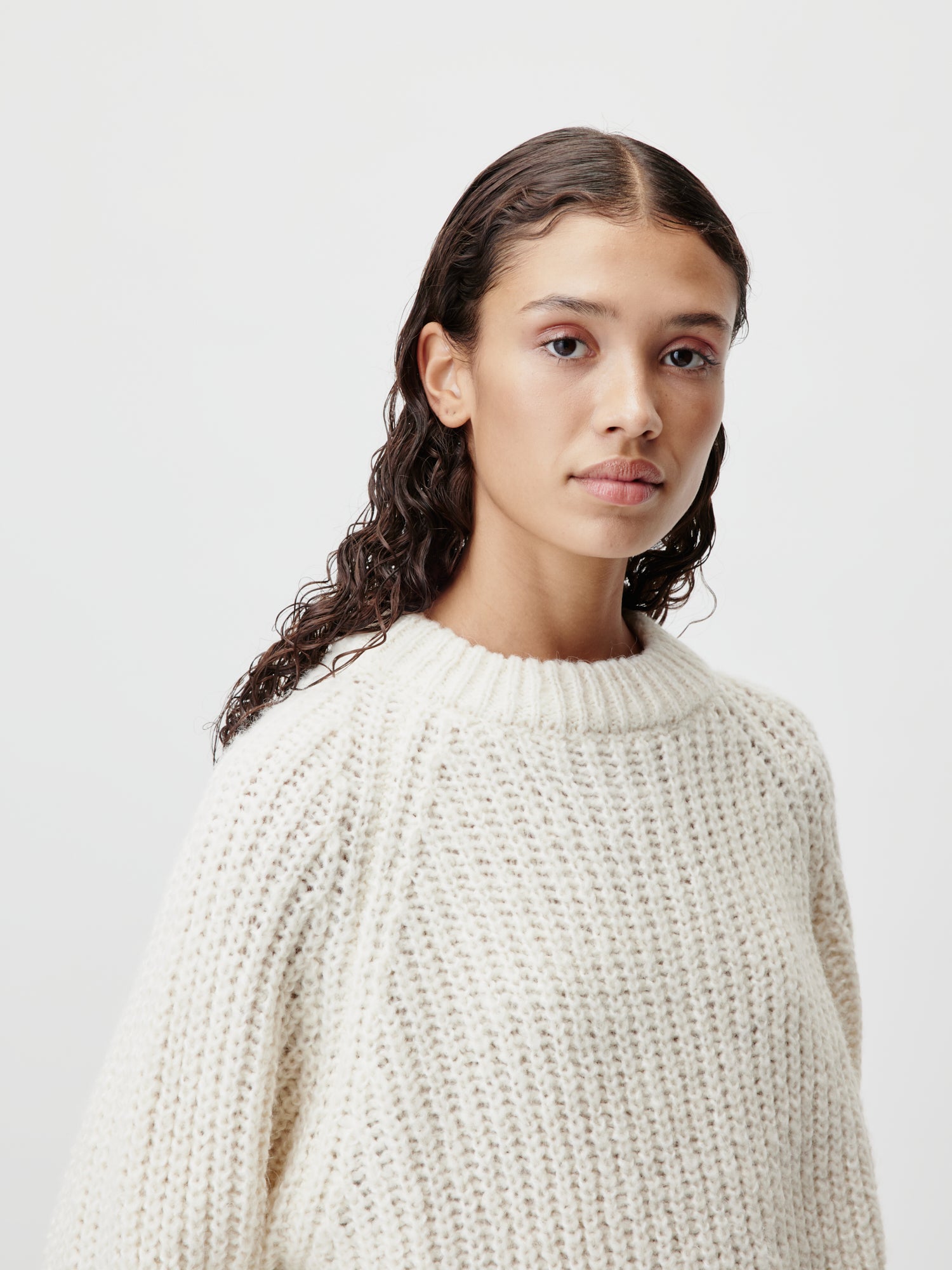 Eine Person mit langem, welligem Haar trägt den Leslie Jumper, einen groben, cremefarbenen Strickpullover von LeGer by Lena Gercke in Größe 36, steht vor einem schlichten weißen Hintergrund und blickt direkt in die Kamera.