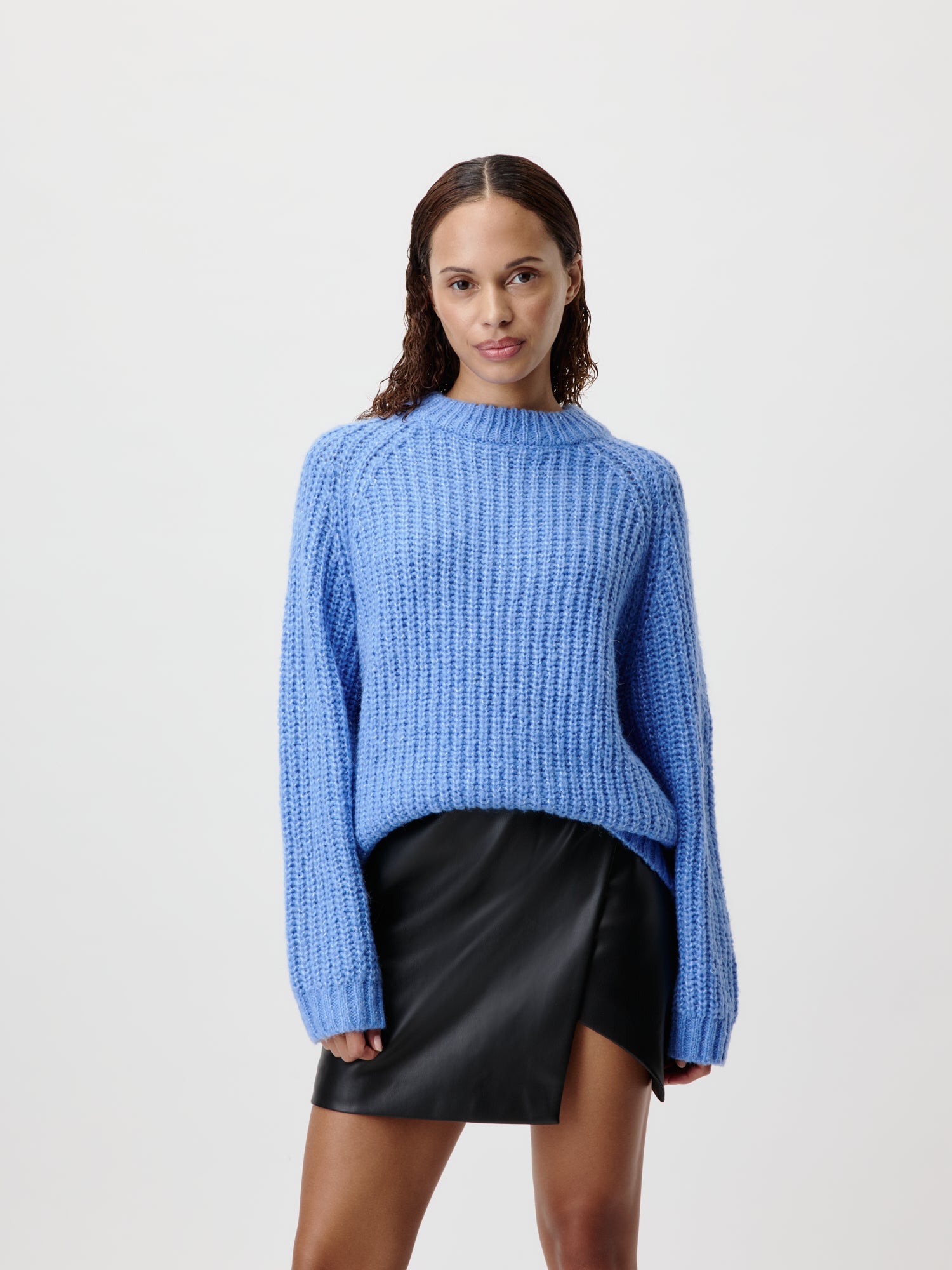 Eine Person mit welligem Haar trägt den Leslie Jumper, einen blauen Strickpullover von LeGer by Lena Gercke mit langen Ärmeln. Dazu trägt sie einen schwarzen Lederrock mit Schlitz vorne in Größe 36. Die Person steht vor einem schlichten weißen Hintergrund und blickt mit neutralem Gesichtsausdruck in die Kamera.