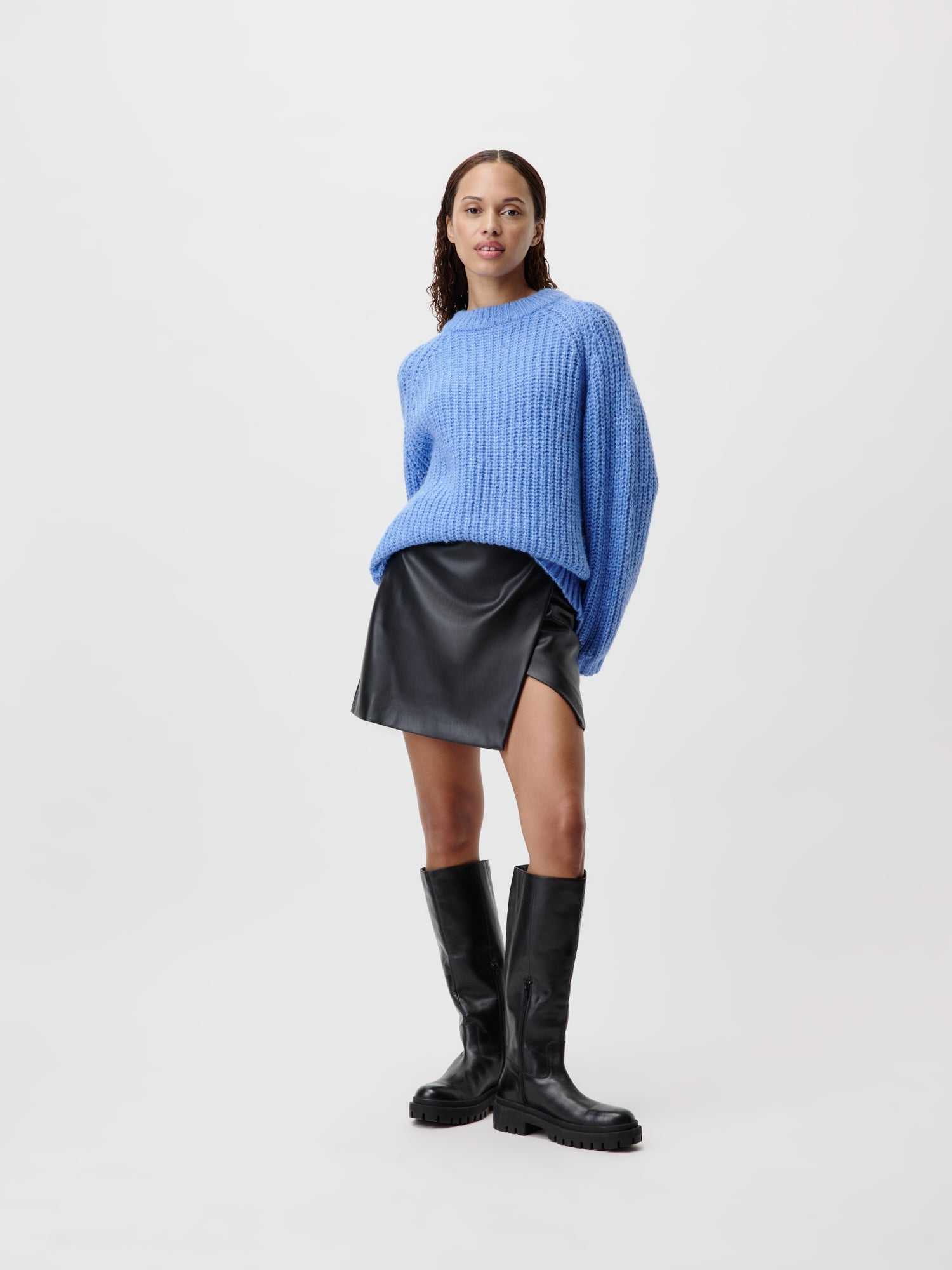 Eine Person posiert selbstbewusst im Leslie Jumper, einem blauen Strickpullover von LeGer by Lena Gercke mit langen Ärmeln. Dazu trägt sie einen schwarzen Lederminirock und kniehohe Stiefel. Sie stehen vor einem schlichten, hellen Hintergrund und tragen Größe 36 für eine perfekte Passform.