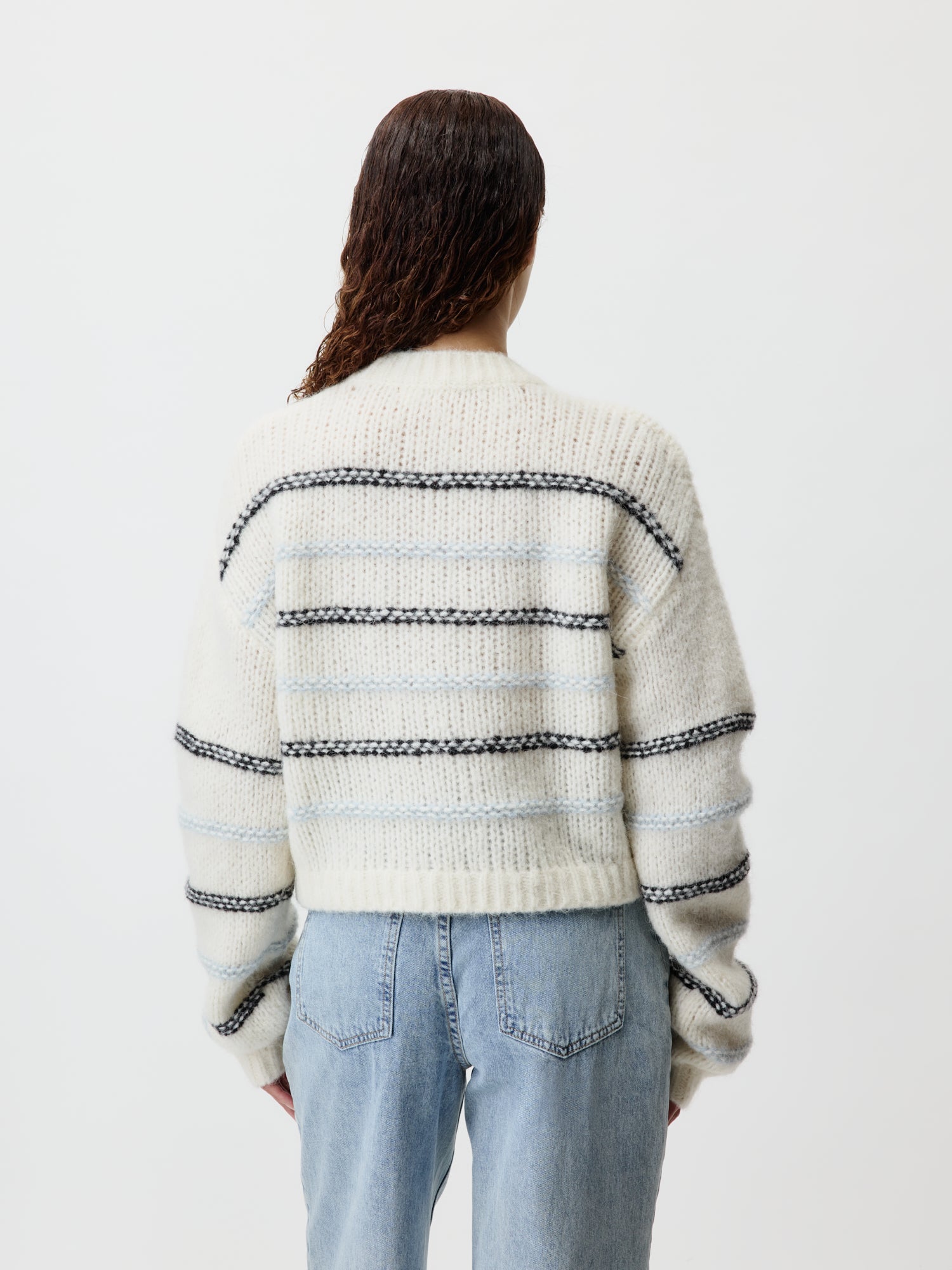 Eine Person steht mit dem Rücken zur Kamera und trägt einen Paulina-Pullover von LeGer by Lena Gercke, ein cremefarbenes, dickes Strickmuster mit horizontalen schwarzen und grauen Streifen. Der Langarm-Stil bietet eine entspannte Passform. Sie hat langes, welliges Haar und trägt hellblaue Jeans vor einem schlichten weißen Hintergrund.