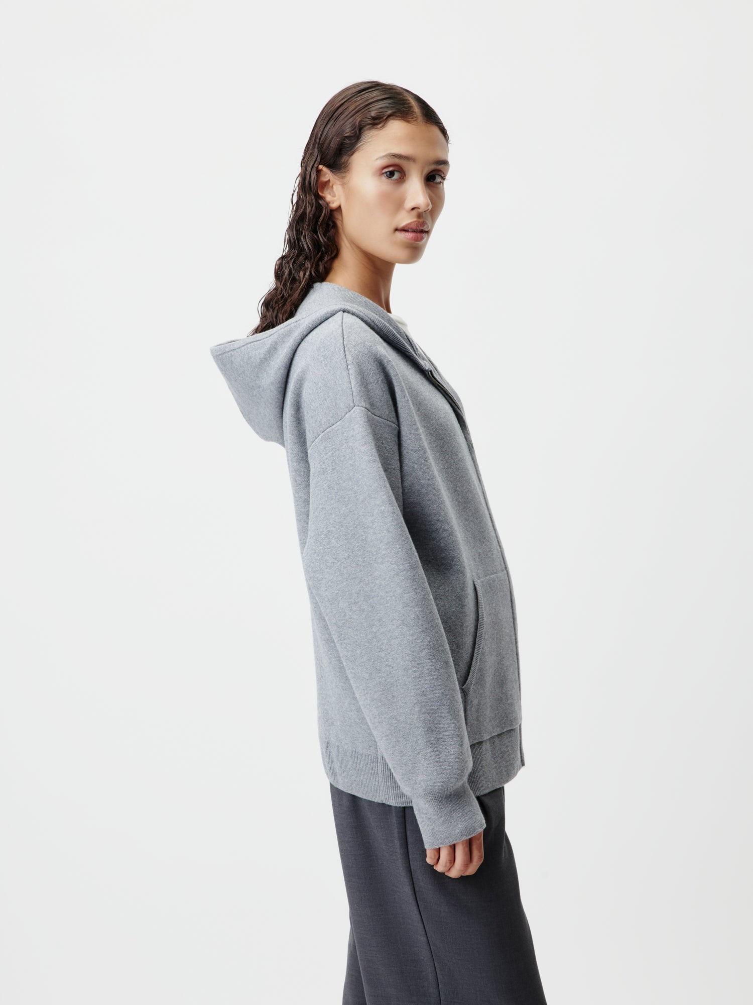 Eine Person mit langen, welligen Haaren trägt den Iriana Knit Hoodie von LeGer by Lena Gercke in Grau und dazu dunkle Hosen. Sie steht seitlich vor einem schlichten weißen Hintergrund, hat einen neutralen Gesichtsausdruck und lässt ihre Hände an den Seiten ruhen.