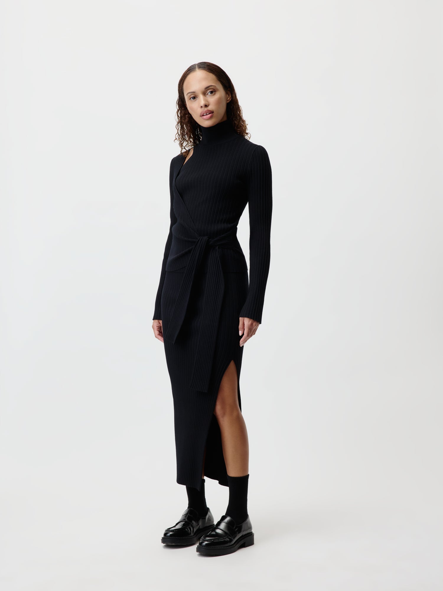 Eine Person trägt den Elenie Jumper von LeGer by Lena Gercke, ein langes, schwarzes, geripptes Kleid mit hohem Kragen und gebundener Taille, das eine figurbetonte Silhouette wunderschön verkörpert. Das Kleid hat einen stilvollen Seitenschlitz. Sie kombinieren es mühelos mit schwarzen Socken und Slippern vor dem schlichten weißen Hintergrund.