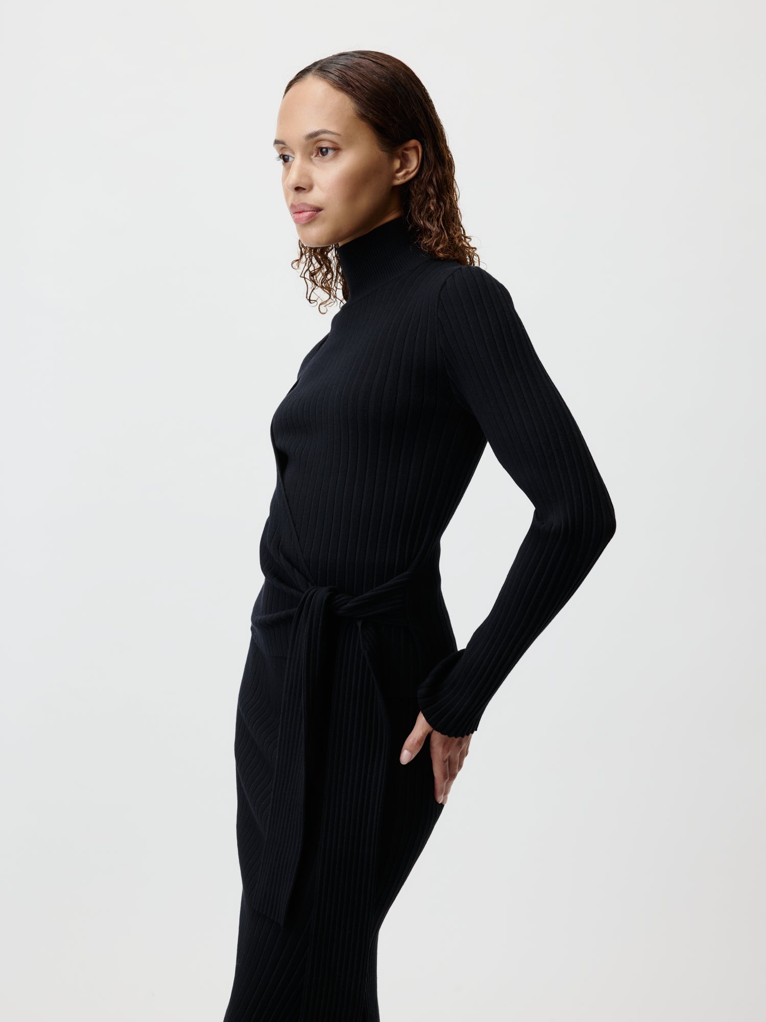 Eine Person mit lockigem Haar präsentiert den Elenie Jumper von LeGer by Lena Gercke, ein schwarzes, geripptes, langärmliges Rollkragenkleid. Das figurbetonte Design hat einen Taillengürtel und fällt normal lang. Sie stehen im Profil vor einem schlichten weißen Hintergrund.