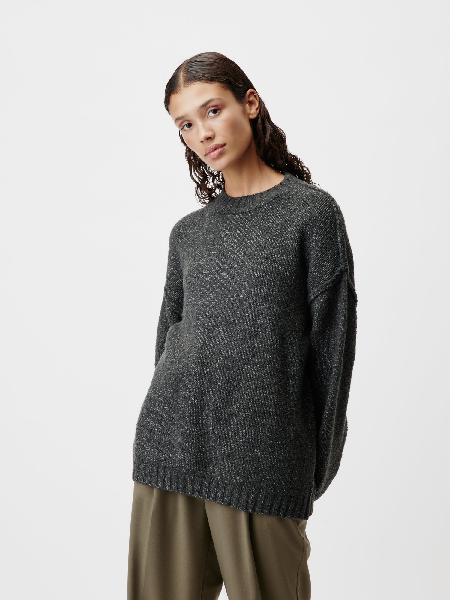 Eine Person mit langen dunklen Haaren trägt einen übergroßen Elsa-Pullover von LeGer by Lena Gercke im langen dunkelgrauen Stil, ergänzt durch locker sitzende Khakihosen vor einem schlichten weißen Hintergrund.