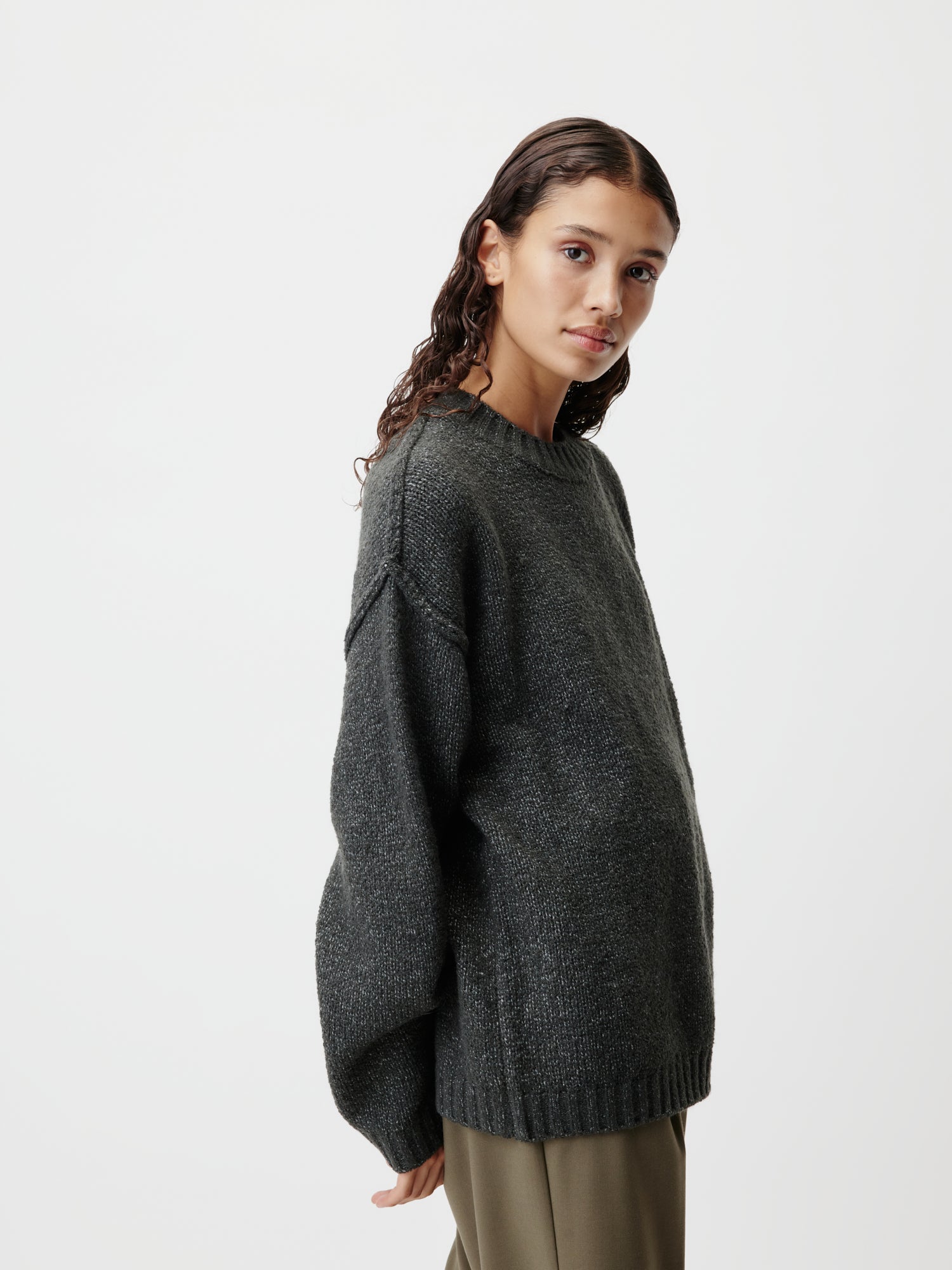 Eine Person mit langen, welligen Haaren trägt einen dunklen, übergroßen Elsa-Pullover von LeGer by Lena Gercke und olivfarbene Hosen. Sie steht seitlich vor einem schlichten, weißen Hintergrund und blickt mit neutralem Gesichtsausdruck in die Kamera – ideal für Ihre nächste Produktinformationsanzeige.