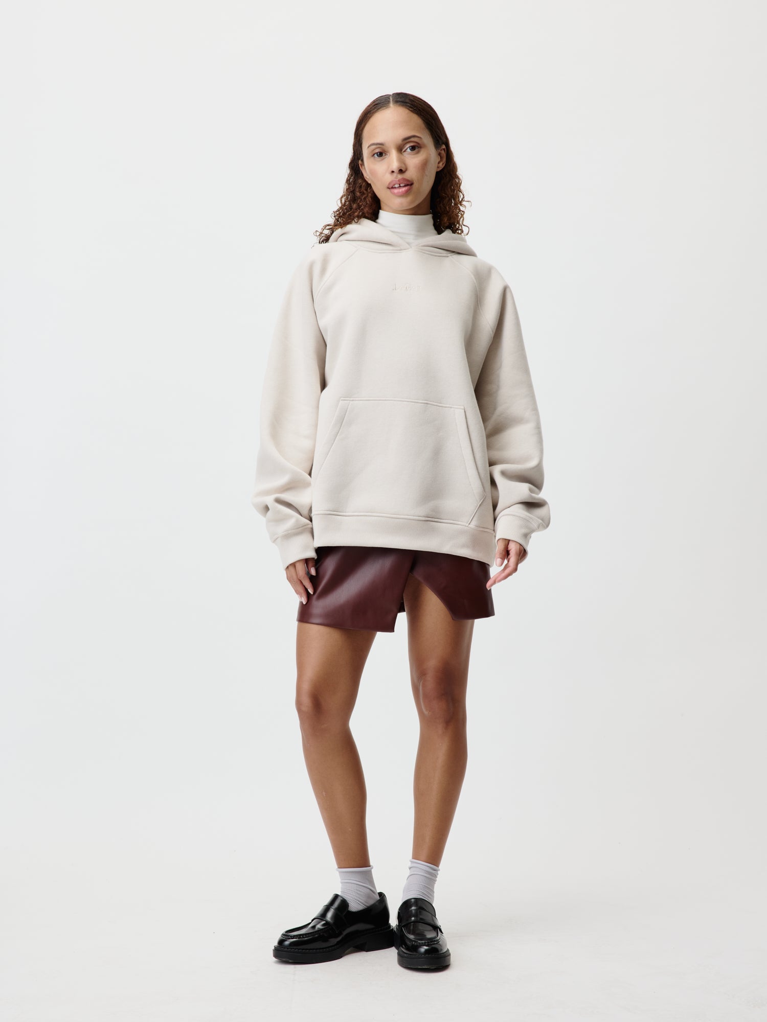 Eine Person mit langen lockigen Haaren trägt einen Naima Hoodie von LeGer by Lena Gercke in Beige, kombiniert mit braunen Shorts, die eine Passform der Größe 36 verkörpern. Sie vervollständigen den Look mit weißen Socken und schwarzen Schuhen vor einem schlichten weißen Hintergrund.