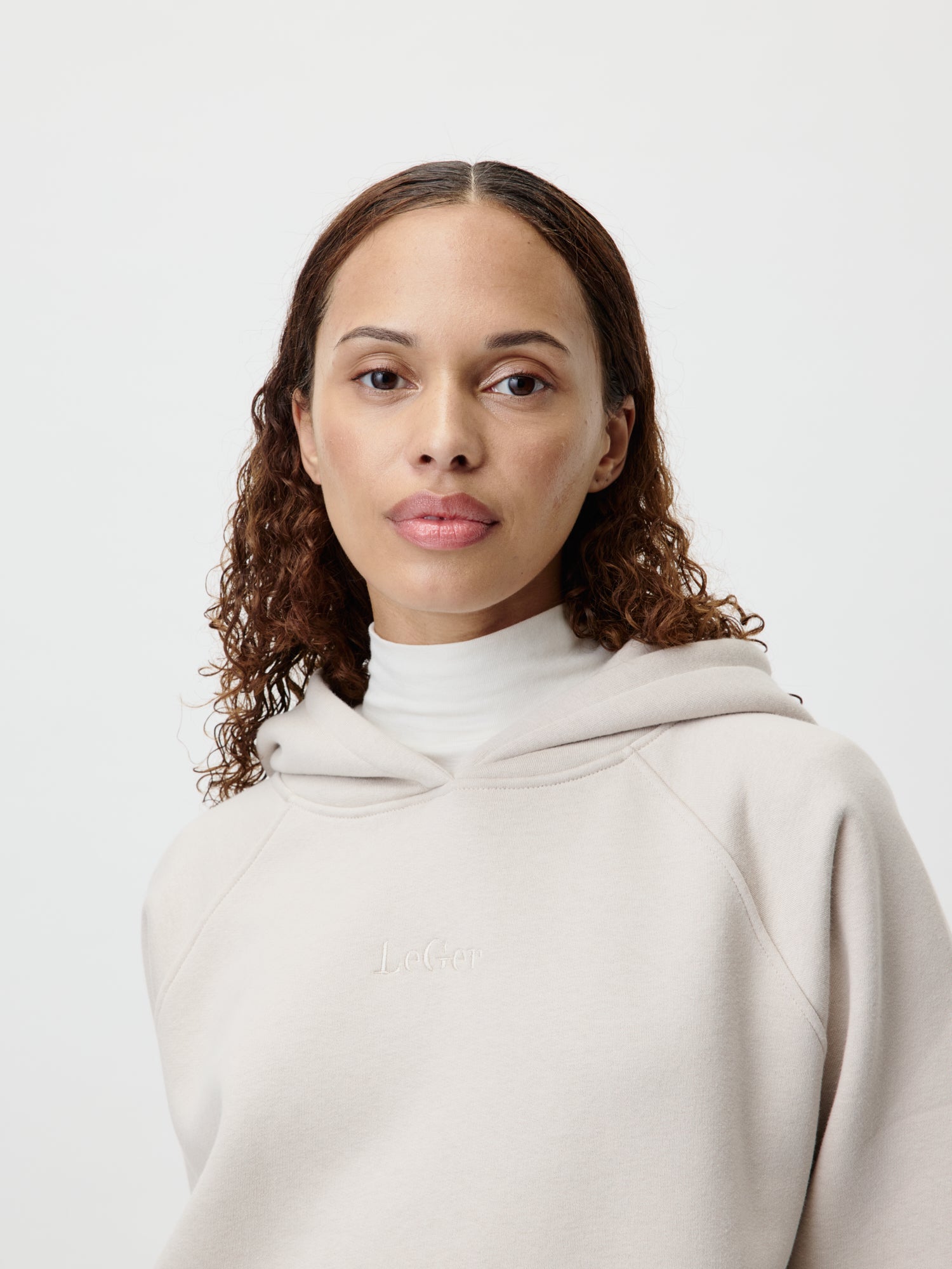 Eine Person mit lockigem braunem Haar trägt einen Naima Hoodie von LeGer by Lena Gercke in Größe 36 in Beige über einem weißen Rollkragenpullover und steht vor einem schlichten hellen Hintergrund. Das Langarm-Design bietet eine stilvolle Passform, während es einen neutralen Ausdruck beibehält.