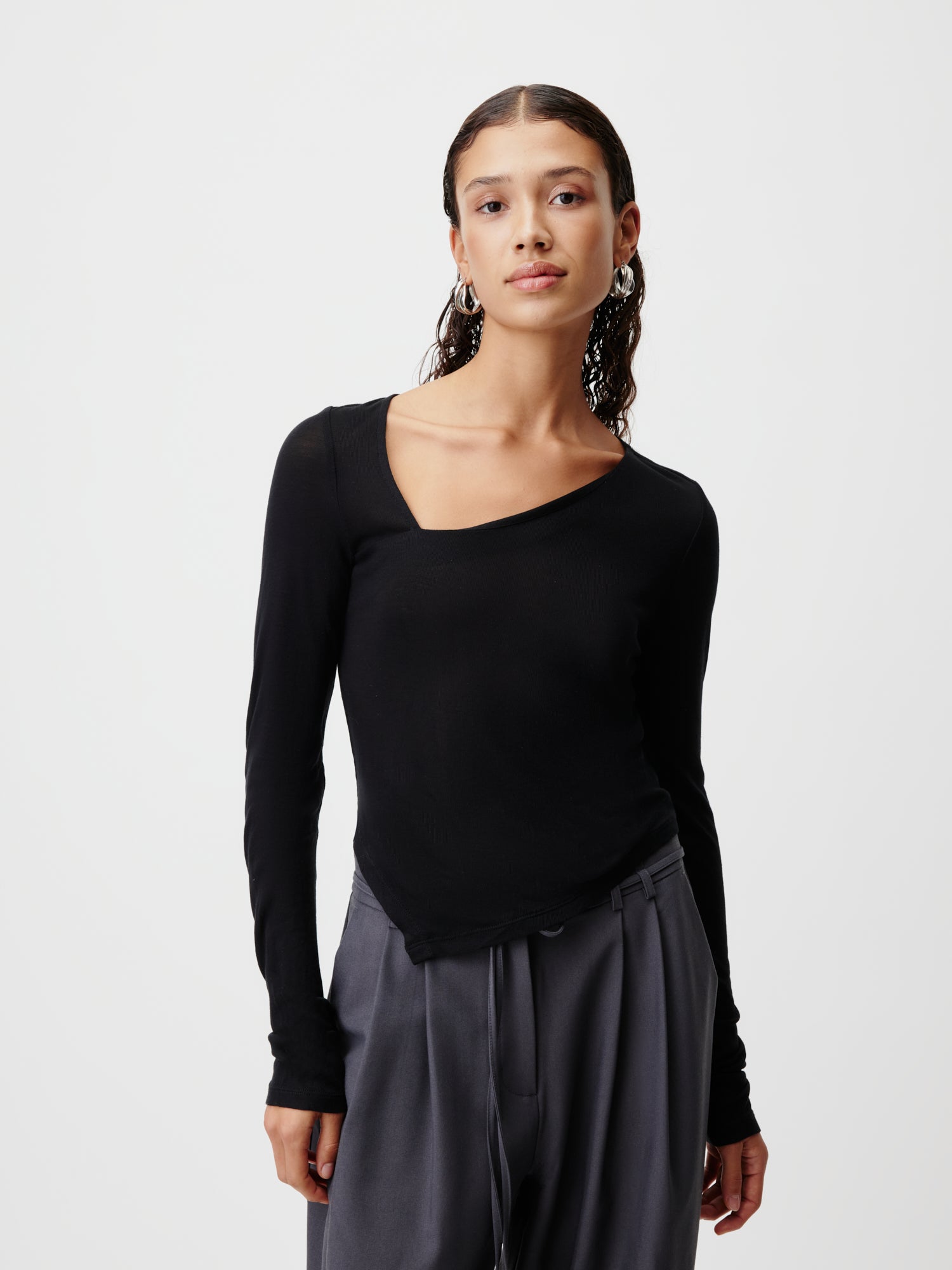 Eine Person mit langen, lockigen Haaren ist elegant gekleidet in einem schwarzen asymmetrischen Gleni Longsleeve von LeGer by Lena Gercke und einer dunklen, schmalen Hose. Sie steht selbstbewusst vor einem schlichten weißen Hintergrund.