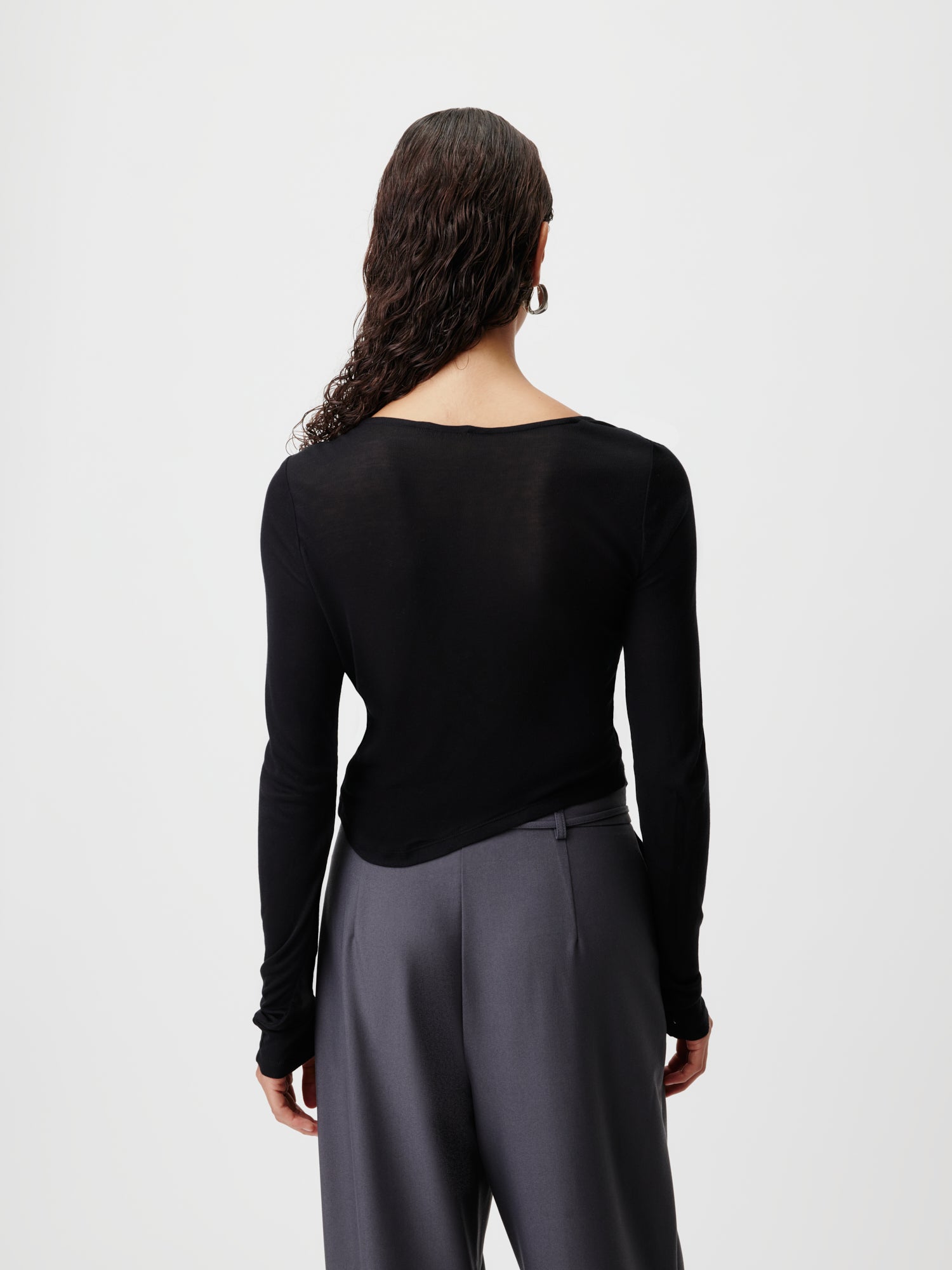 Von hinten ist eine Person mit langen, lockigen Haaren zu sehen, die das Gleni Longsleeve von LeGer by Lena Gercke in einer schwarz-grauen Hose trägt. Der Hintergrund bleibt schlicht weiß.