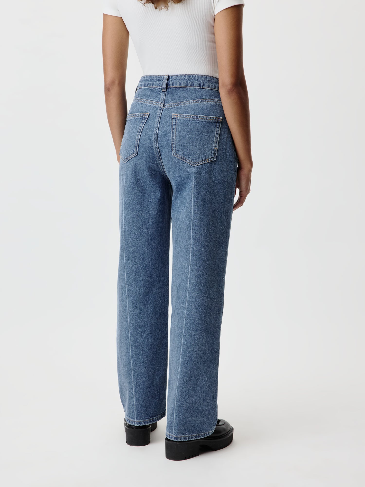 Rückansicht einer Person in Cathlin Pants von LeGer by Lena Gercke, Größe 36, aus hoch tailliertem blauem Denim mit zwei Gesäßtaschen und weitem Lang-/Maxi-Bein, kombiniert mit einem weißen Oberteil und schwarzen Schuhen, vor einem einfarbigen Hintergrund.