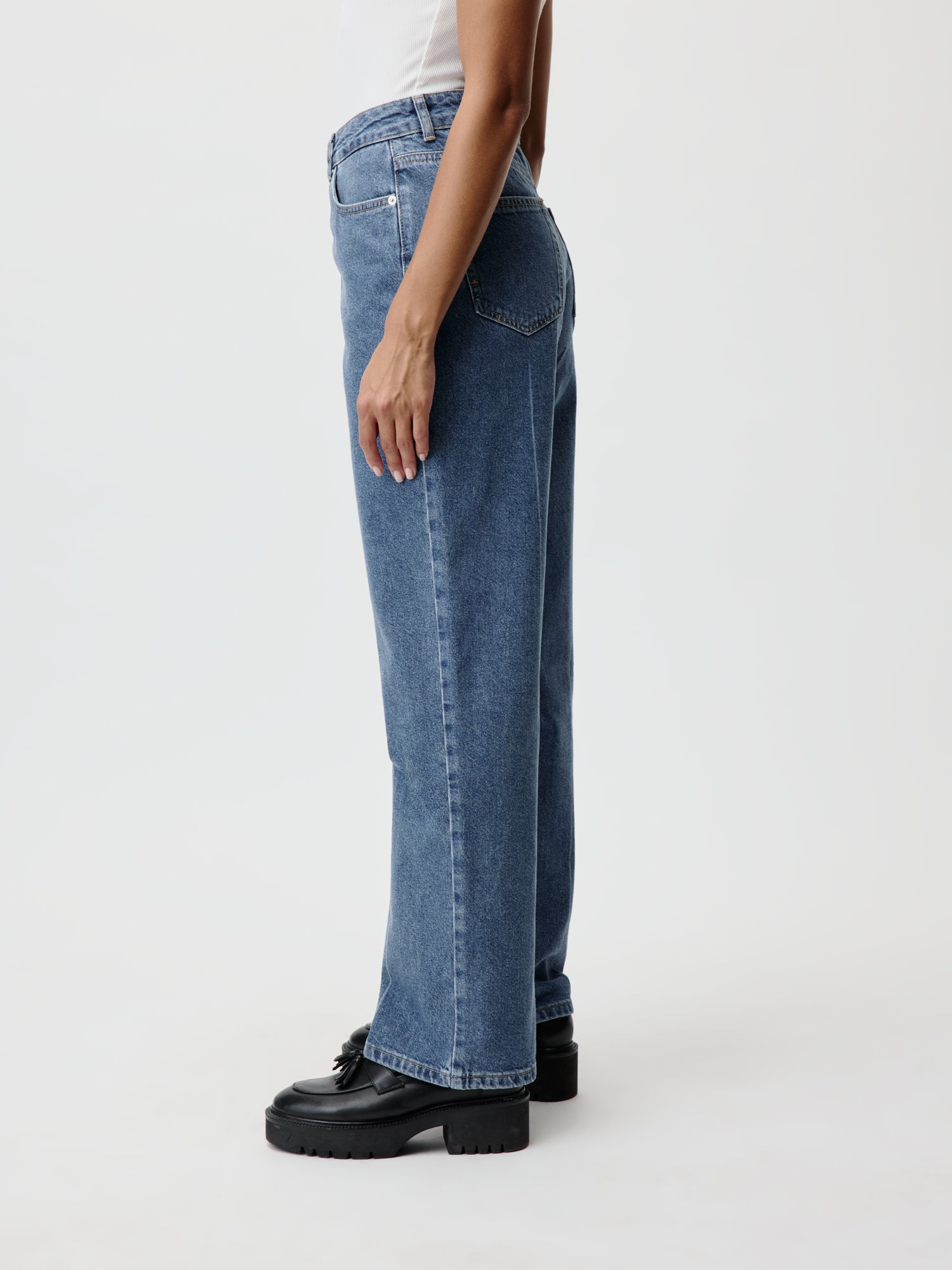 Das Modell in Größe 36 präsentiert sich in einem stylischen weißen ärmellosen Top kombiniert mit der Cathlin Hose von LeGer by Lena Gercke, einem locker sitzenden Bluejeans-Design mit geradem Bein. Der schicke Look wird durch schwarze Plateauschuhe komplettiert, die ihn vor dem schlichten weißen Hintergrund hervorstechen lassen.