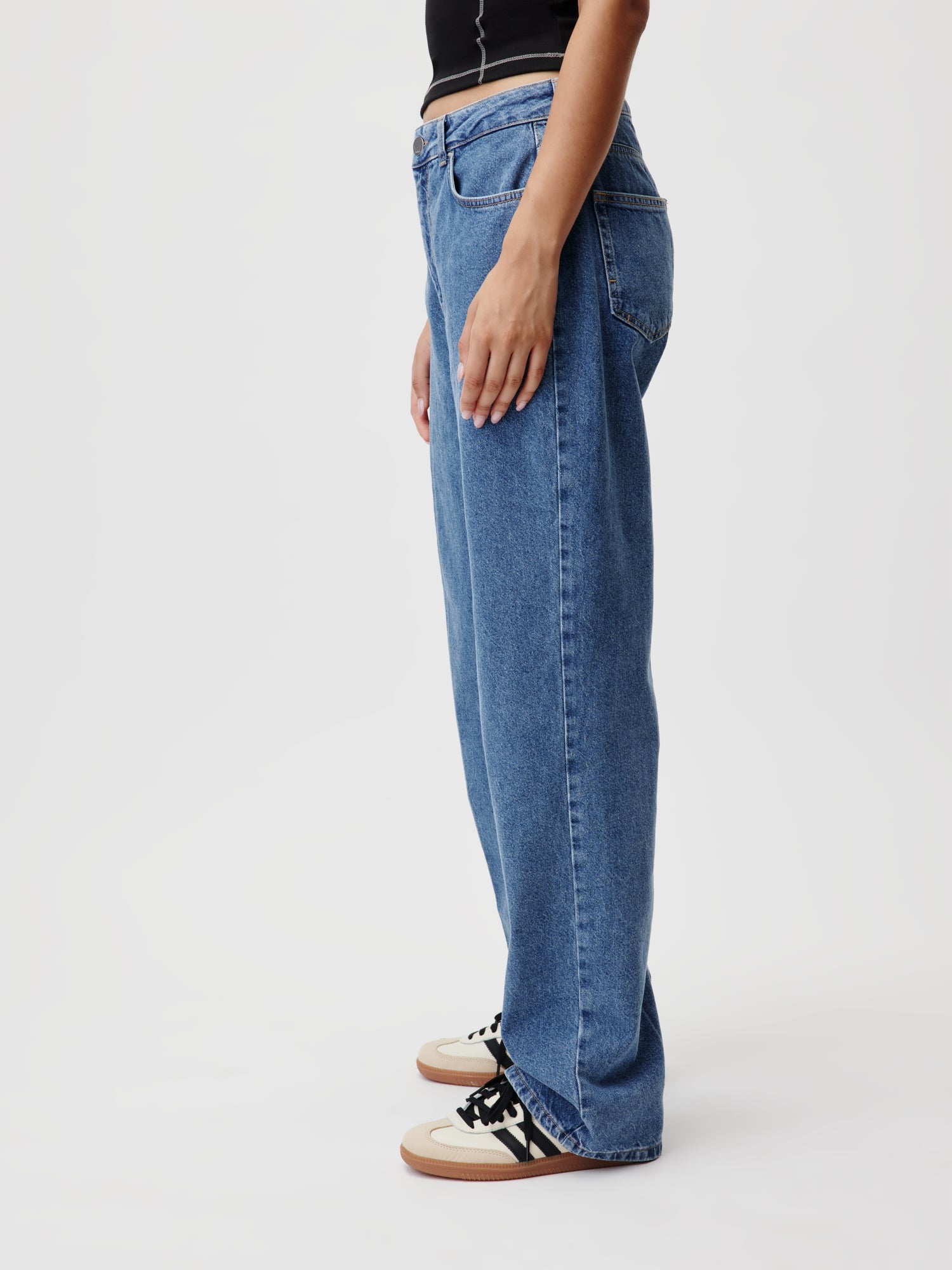 Eine Person steht seitlich und trägt locker sitzende blaue Cathlin Pants Tall von LeGer by Lena Gercke und ein schwarzes Oberteil, perfekt für Größe 36. Ihre Füße stecken in weißen Sneakers mit schwarzen Details und hellbraunen Sohlen. Der schlichte weiße Hintergrund betont die Körpergröße des Models von 1,77 Metern.