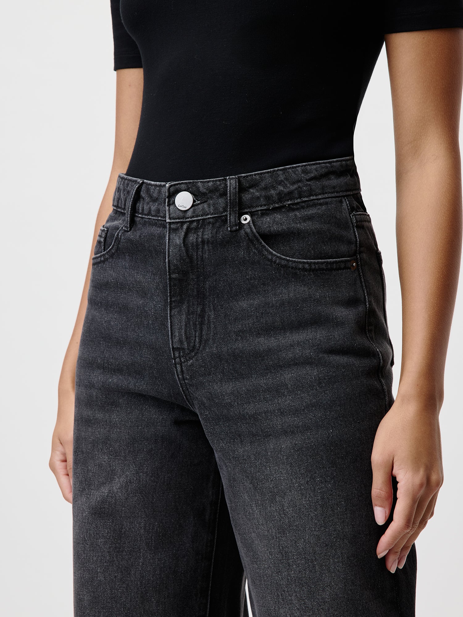 Eine Person in einem schwarzen kurzärmeligen Oberteil und hoch taillierten Marin Pants von LeGer by Lena Gercke steht mit entspannten Händen vor einem schlichten weißen Hintergrund und strahlt zeitlose Eleganz aus. Für weitere Produktinformationen zu Länge und Passform der Marin Pants können Sie sich gerne an uns wenden.