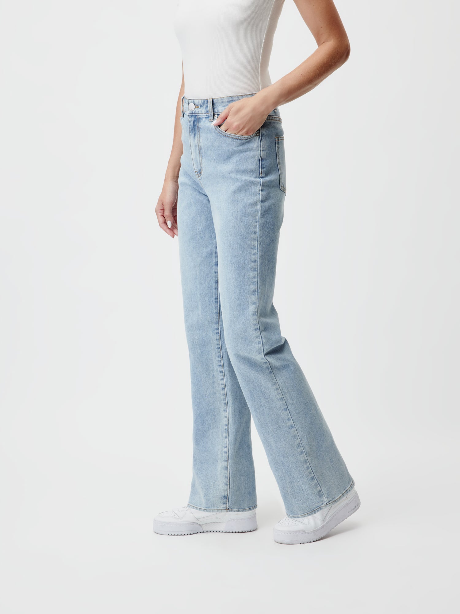 Eine Person trägt die Falda Pants Tall von LeGer by Lena Gercke in Größe 38, mit entspannter Passform und hellblauem Denim-Look, und posiert vor einem schlichten weißen Hintergrund. Sie trägt außerdem ein weißes Tanktop mit passenden weißen Sneakers und hat lässig eine Hand in der Tasche der Jeans vergraben.