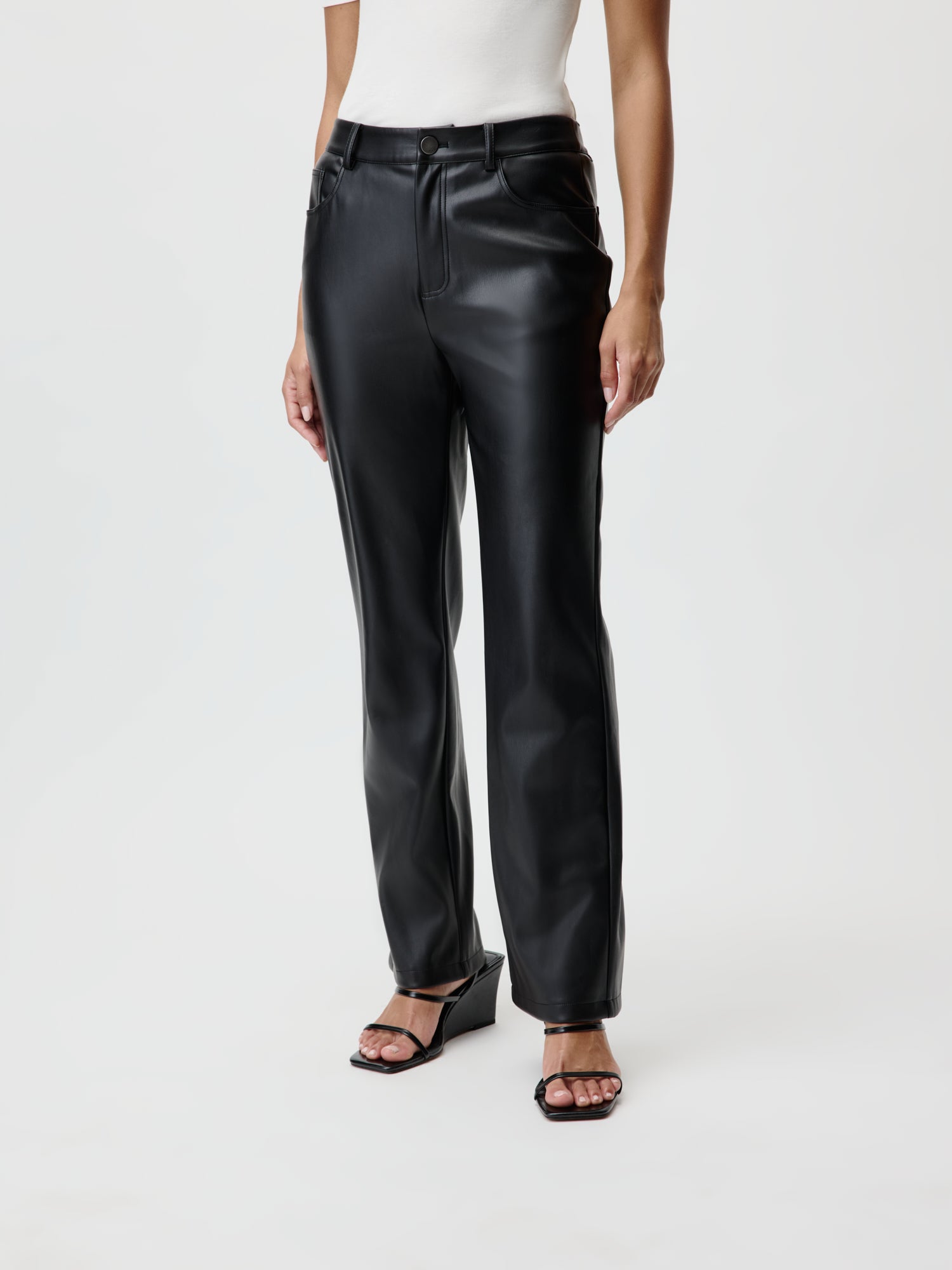 Eine Person, die ein weißes ärmelloses Oberteil und Naomi Pants aus schwarzem Leder von LeGer by Lena Gercke trägt, steht mit den Händen an den Seiten. Der elegante Look wird durch schwarze offene Pumps ergänzt. Trotz der Länge des Ensembles bleibt der minimalistische Hintergrund schlicht weiß, sodass jedes Element hervorsticht.