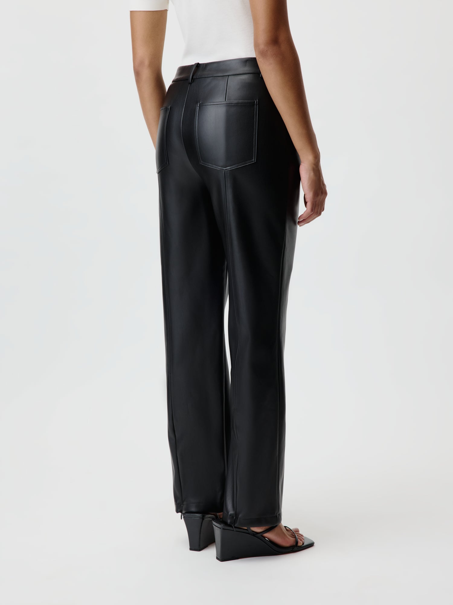 Eine Person trägt die Naomi Pants aus schwarzem Leder von LeGer by Lena Gercke, kombiniert mit einem schicken weißen Oberteil, und steht mit dem Rücken zur Kamera. Ihr Look wird durch stylische schwarze Keilsandalen vor einem schlichten, hellen Hintergrund vervollständigt, der die Lang-Eleganz der Szene unterstreicht.