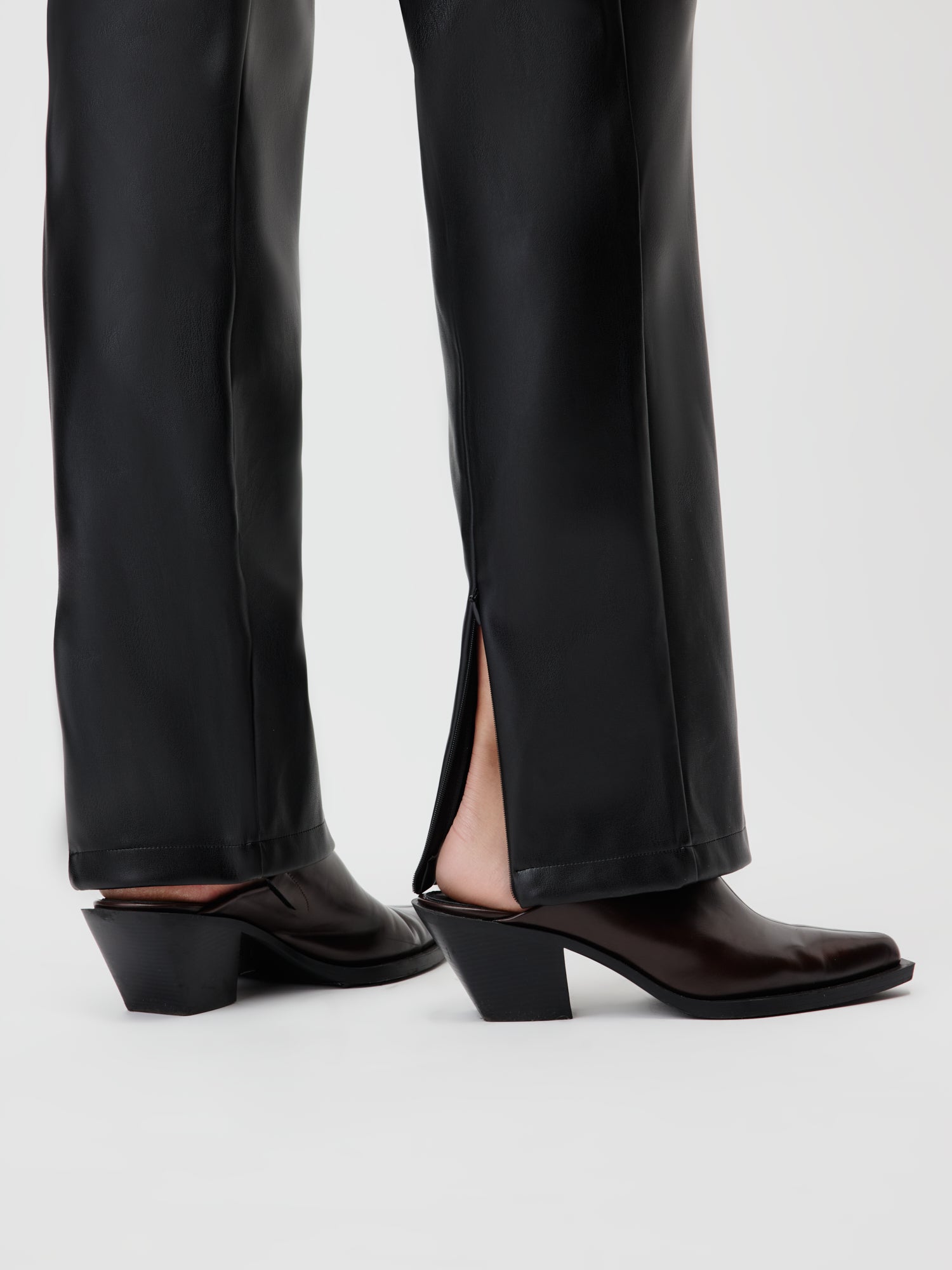 Nahaufnahme einer Person, die die Naomi Pants Tall von LeGer by Lena Gercke in Größe 38 mit Schlitzsaum und schwarzen Schuhen mit Absatz trägt. Der Fokus liegt auf den Unterschenkeln und Füßen und zeigt das stilvolle Design und die Länge dieser schwarzen Lederhose vor einem hellen Hintergrund.