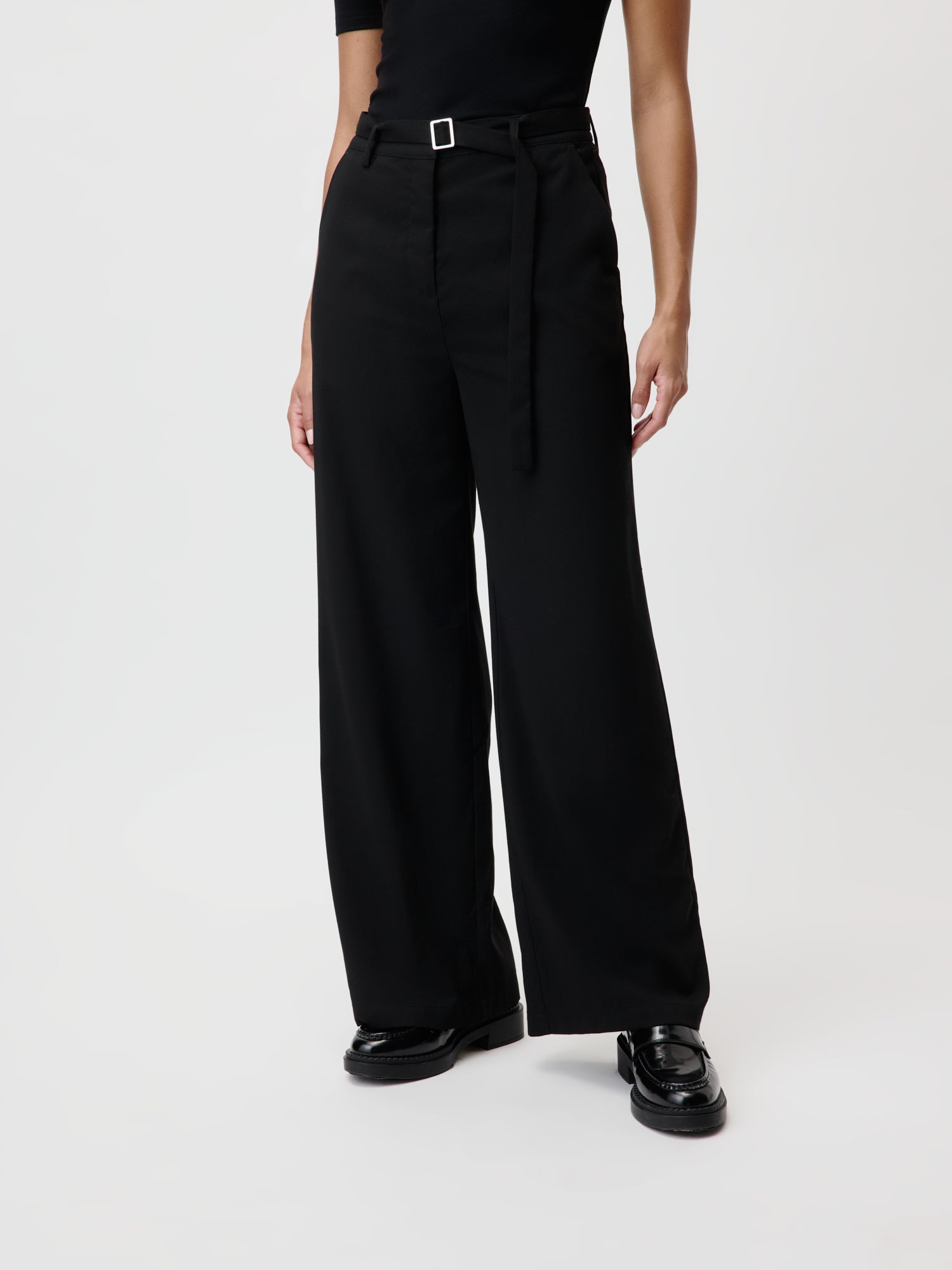 Eine Person trägt die Jila Pants von LeGer by Lena Gercke, die hoch tailliert und schwarz sind und eine Silhouette mit weitem Bein haben, die Länge ausstrahlt. Das Outfit wird durch einen passenden Gürtel und glänzende schwarze Schuhe ergänzt, die die lange Silhouette vor einem schlichten weißen Hintergrund perfekt hervorheben.