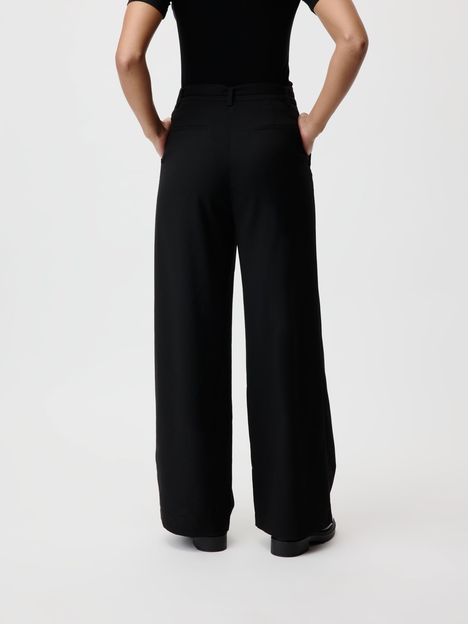 Rückansicht einer Person, die ein schickes schwarzes Oberteil und die Jila Pants von LeGer by Lena Gercke trägt, mit weitem Bein und Maxi-Länge. Sie steht mit den Händen in den Taschen vor einem schlichten weißen Hintergrund und präsentiert ein lässiges, monochromes Ensemble.