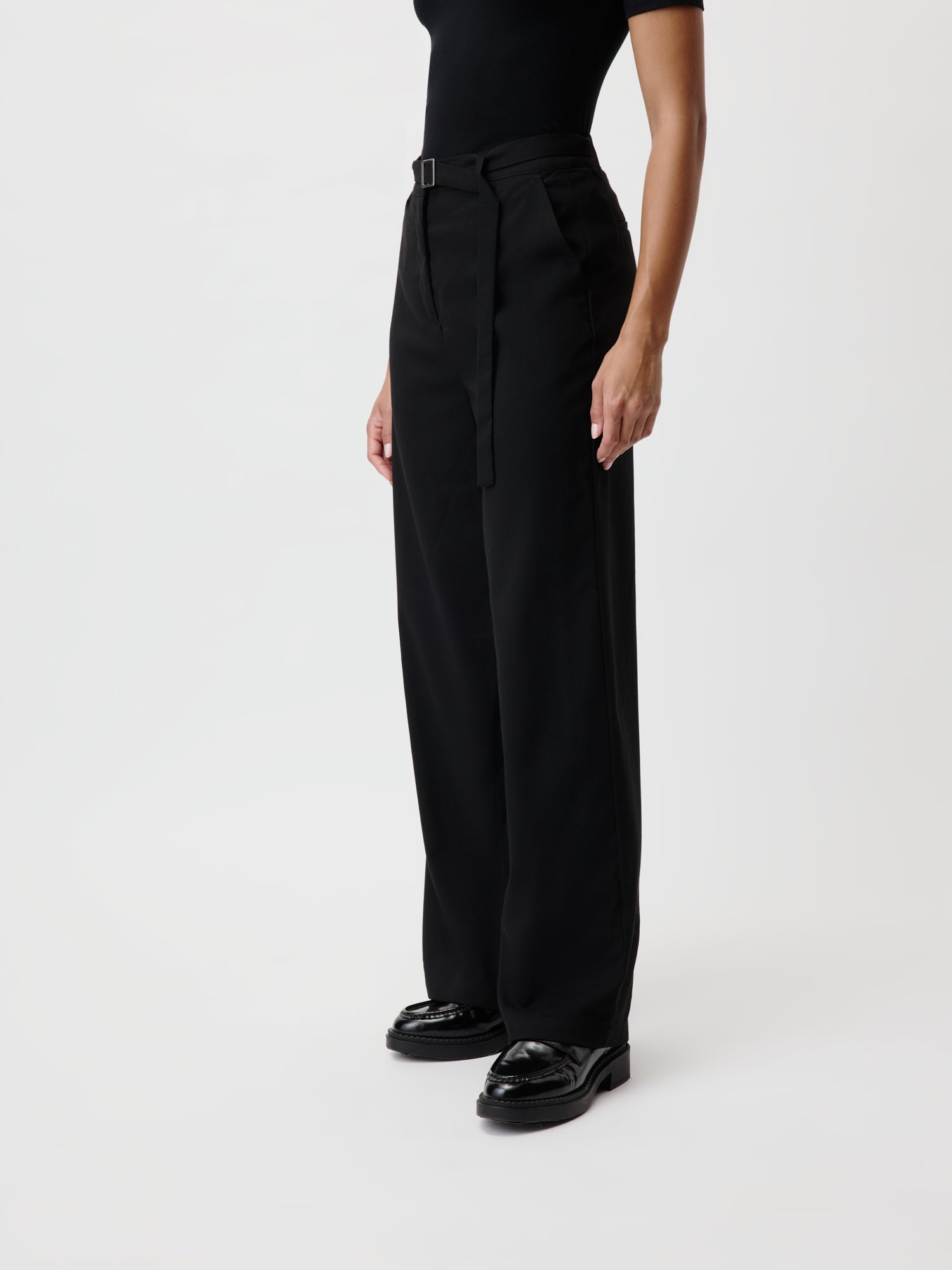 Eine Person trägt ein schwarzes Oberteil und die Jila Pants, eine hoch taillierte schwarze Hose mit weitem Bein und Gürtel von LeGer by Lena Gercke, dazu glänzende schwarze Schuhe. Die Länge des Outfits verleiht vor dem weißen Hintergrund eine elegante Note.