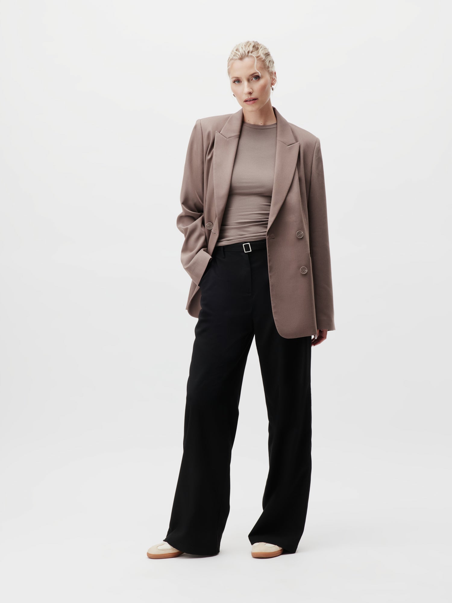 Eine Person steht selbstbewusst vor einem schlichten Hintergrund und betont ihre längliche Silhouette in den schwarzen Jila Pants Tall von LeGer by Lena Gercke. Sie trägt einen taupefarbenen Blazer über einem passenden Oberteil, hat eine Hand in der Tasche und kombiniert das Outfit mit beigen Schuhen.