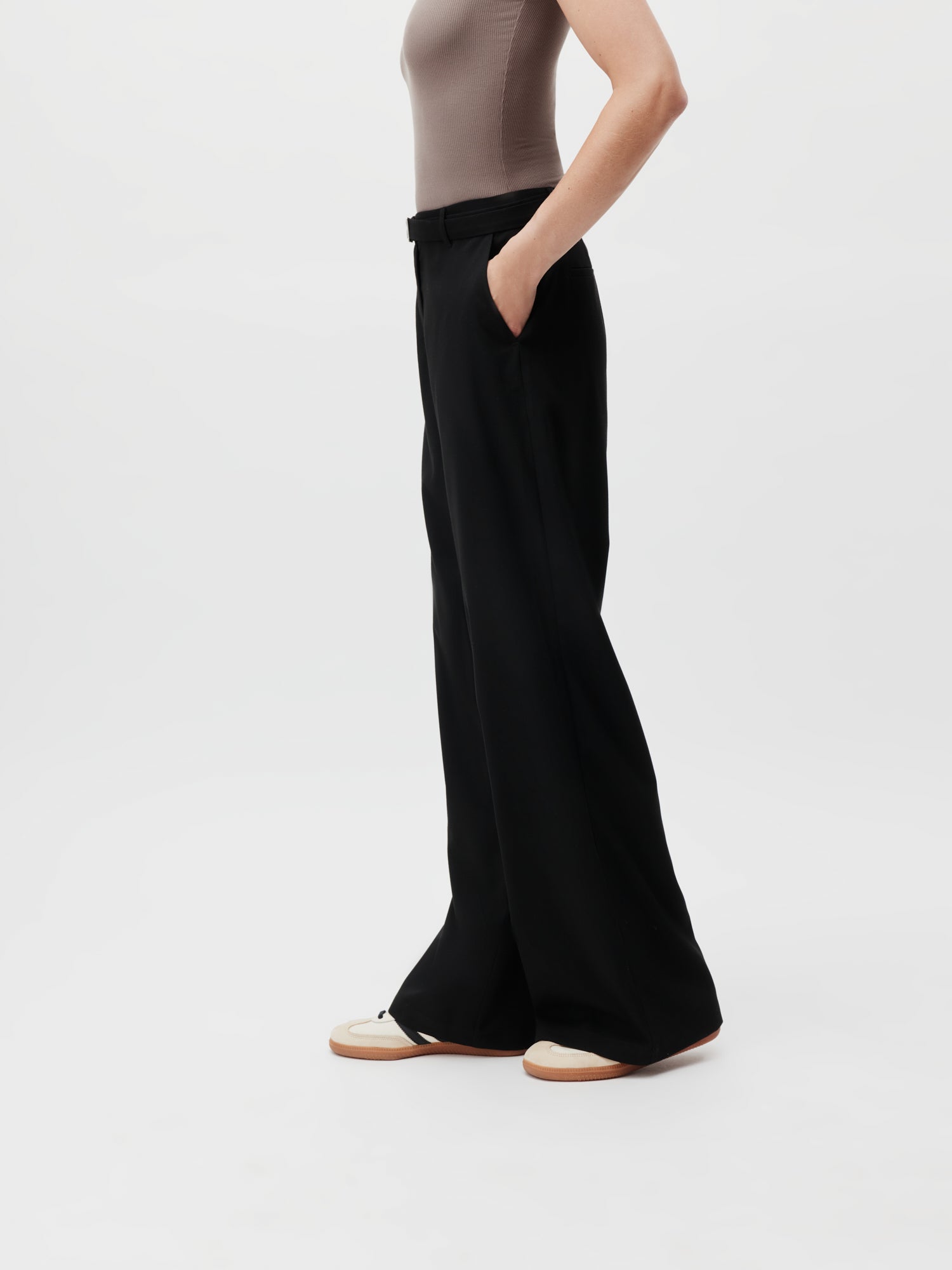 Die Person trägt ein ärmelloses beiges Top und schwarze Jila Pants Tall von LeGer by Lena Gercke. Sie steht vor einem schlichten Hintergrund und strahlt mit ihrer schlanken Silhouette mühelos Stil aus. Ihre rechte Hand ruht in der Tasche, während weiße und hellbraune Schuhe den Look ergänzen. Der Fokus liegt auf dem eleganten Maxi-Outfit.