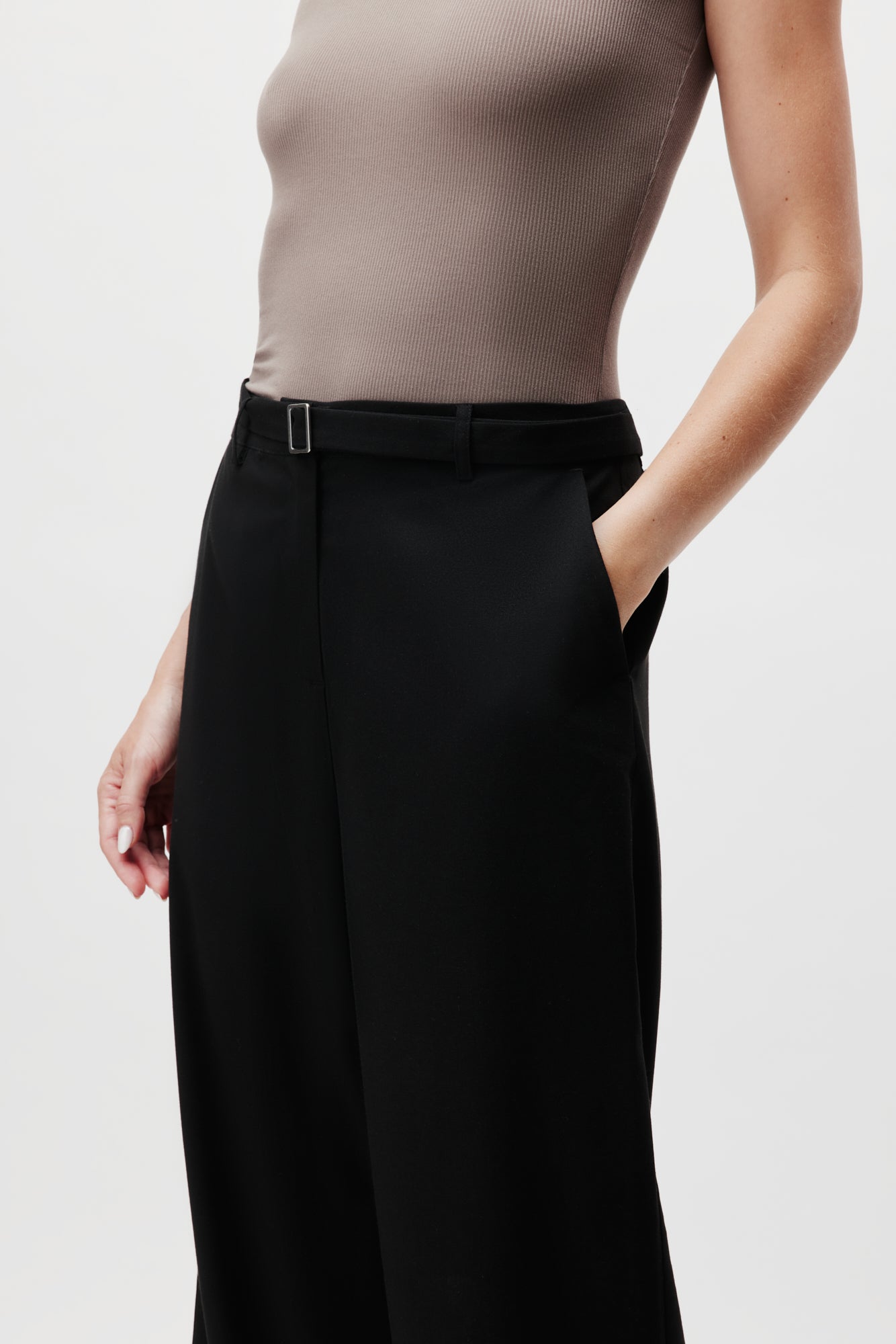 Hier werden die Jila Pants Tall von LeGer by Lena Gercke präsentiert, kombiniert mit einem ärmellosen taupefarbenen Oberteil, um eine Mischung aus Eleganz und Schlichtheit zu schaffen. Die Hand des Models ruht lässig in einer Tasche, was das schlanke Design vor einem schlichten Hintergrund betont und die Essenz des minimalistischen Stils von LeGer einfängt.