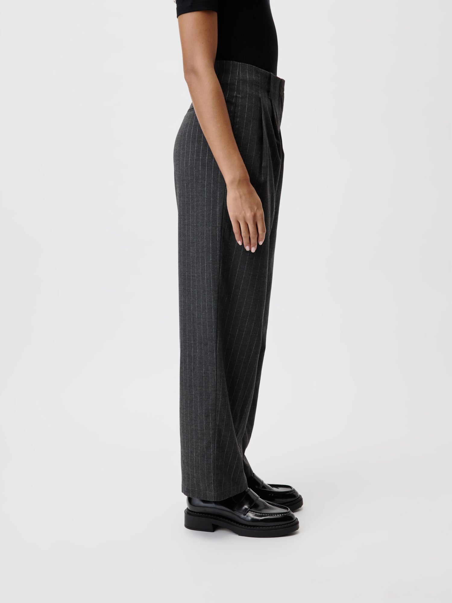 Eine Person steht im Seitenprofil und trägt ein schwarzes Hemd und hoch taillierte, graue Delphine-Hosen mit Nadelstreifen von LeGer by Lena Gercke, kombiniert mit schwarzen Slippern. Dieser minimalistische Look spiegelt die Eleganz eines Lang-Designs vor dem schlichten weißen Hintergrund wider.