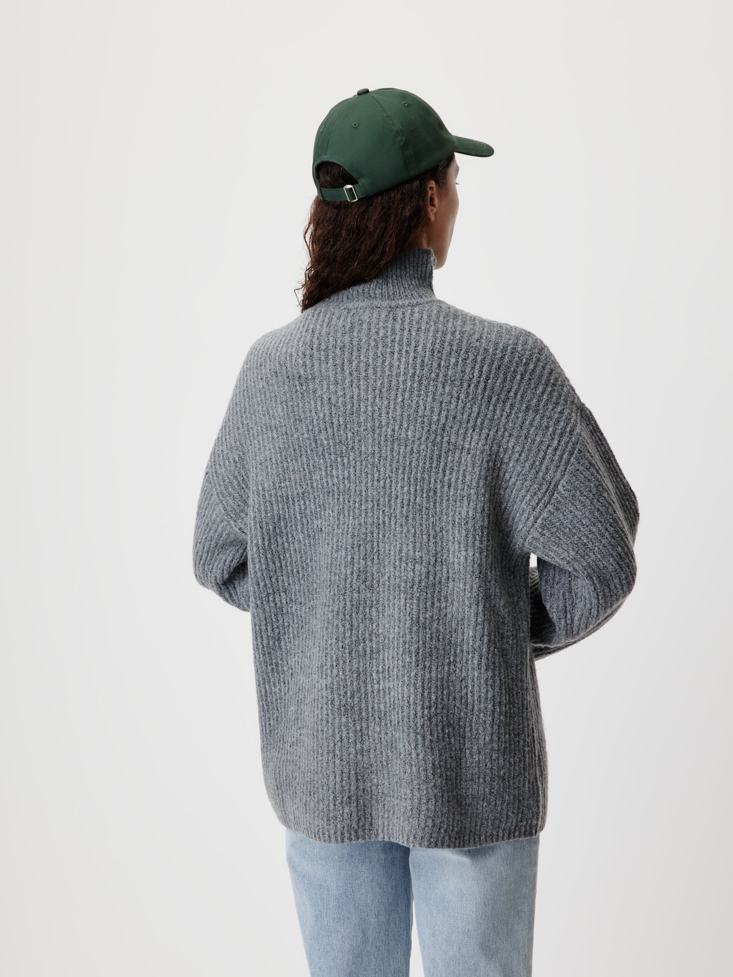 Eine Person mit langen Haaren, einer grünen Baseballkappe und dem Lina Jumper von LeGer by Lena Gercke – einem grauen Rippenpullover in normaler Länge und Langarm-Passform – blickt von der Kamera weg. Sie trägt außerdem hellblaue Jeans vor einem schlichten weißen Hintergrund.