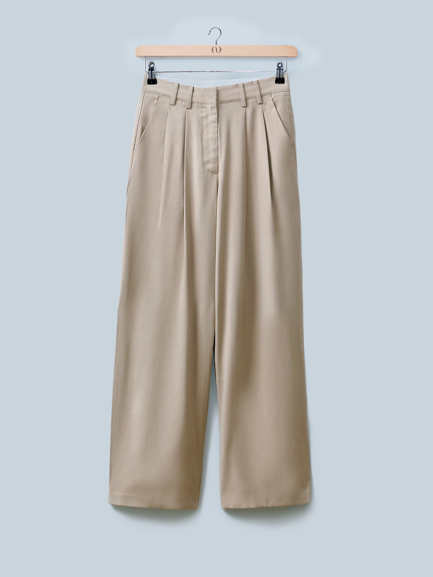 Die Elin Pants von LeGer by Lena Gercke sind beige, weit geschnittene Anzughose mit Bügelfalten und hoher Taille, die auf einem Holzbügel vor hellblauem Hintergrund präsentiert wird.