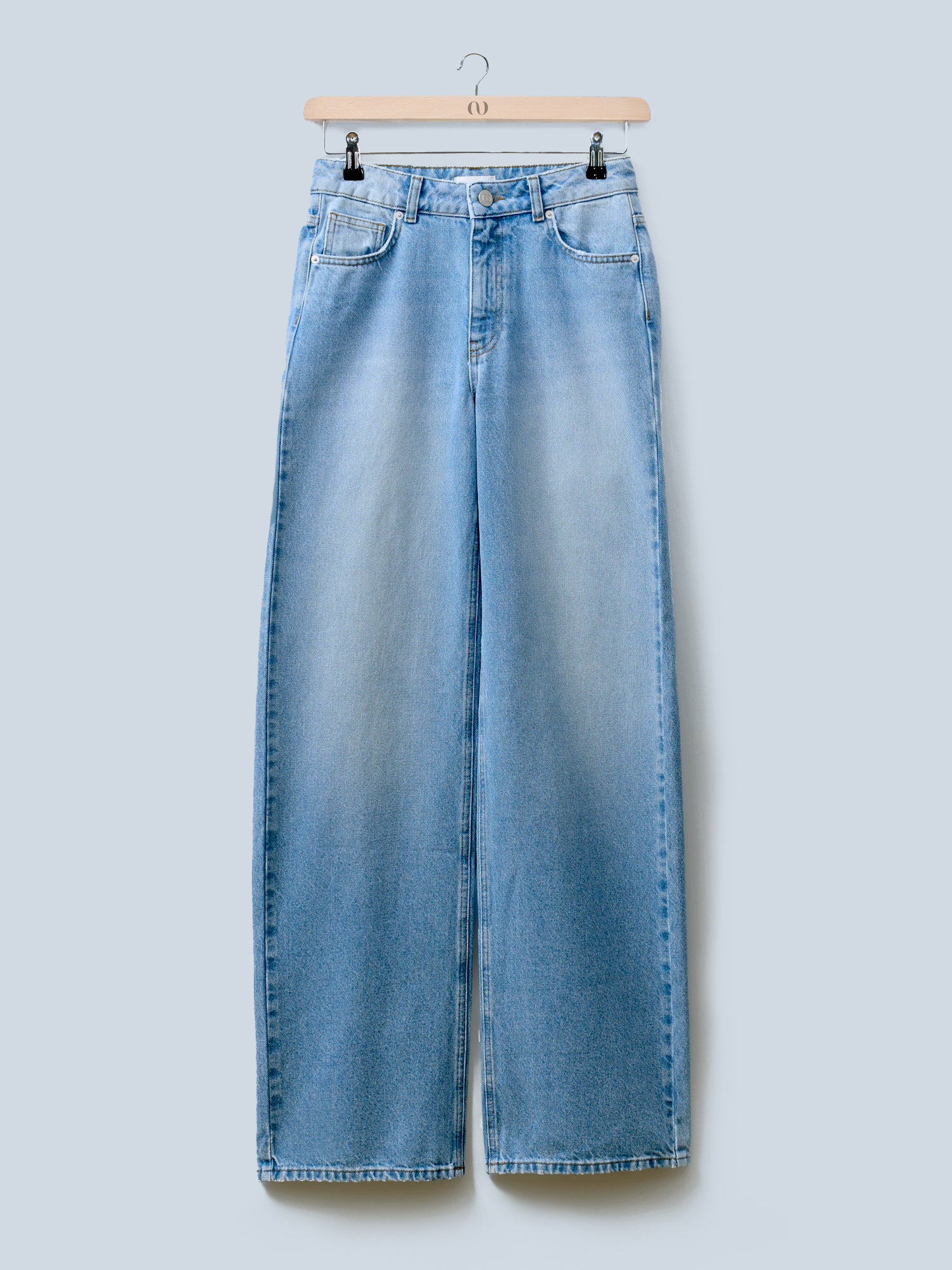 Die Rhonda Pants von LeGer by Lena Gercke, eine hoch taillierte, hellblaue Jeans mit weitem Bein, Knopfverschluss und Vordertaschen, hängt auf einem hölzernen Kleiderbügel vor einem schlichten hellblauen Hintergrund. Die Jeans hat ein leicht verwaschenes Finish.