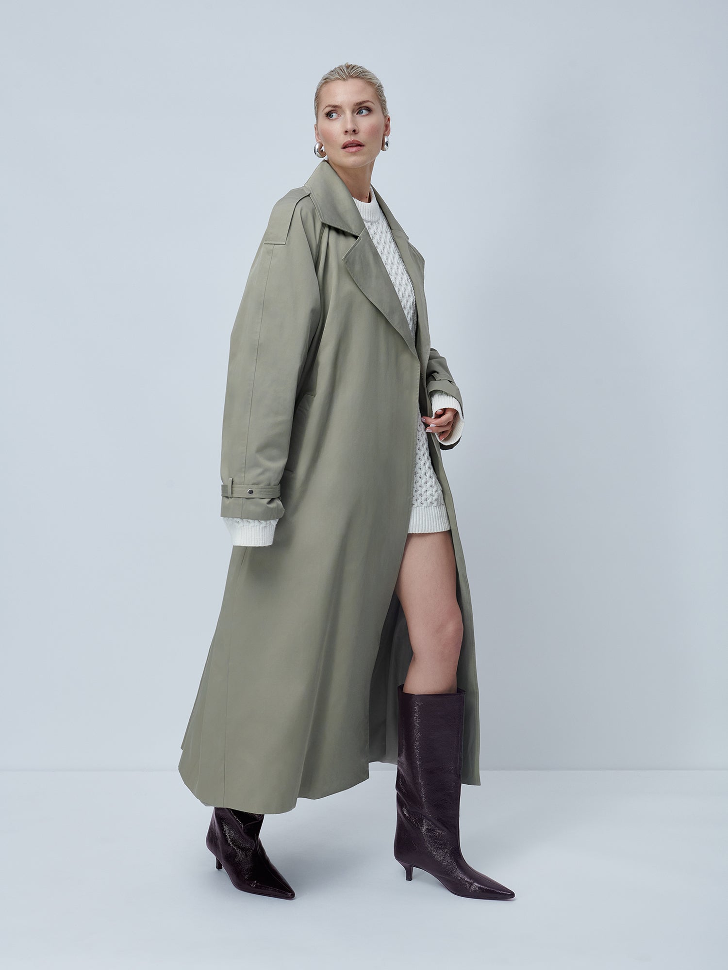 Eine Frau im Elena Coat von LeGer by Lena Gercke, einem langen, salbeigrünen Stück mit Reverskragen, über einem weißen Zopfmuster-Outfit, gepaart mit dunklen kniehohen Stiefeln. Sie steht vor einem minimalistischen grauen Hintergrund mit den Händen in den Taschen, blickt leicht zur Seite und strahlt zeitloses klassisches Design aus.